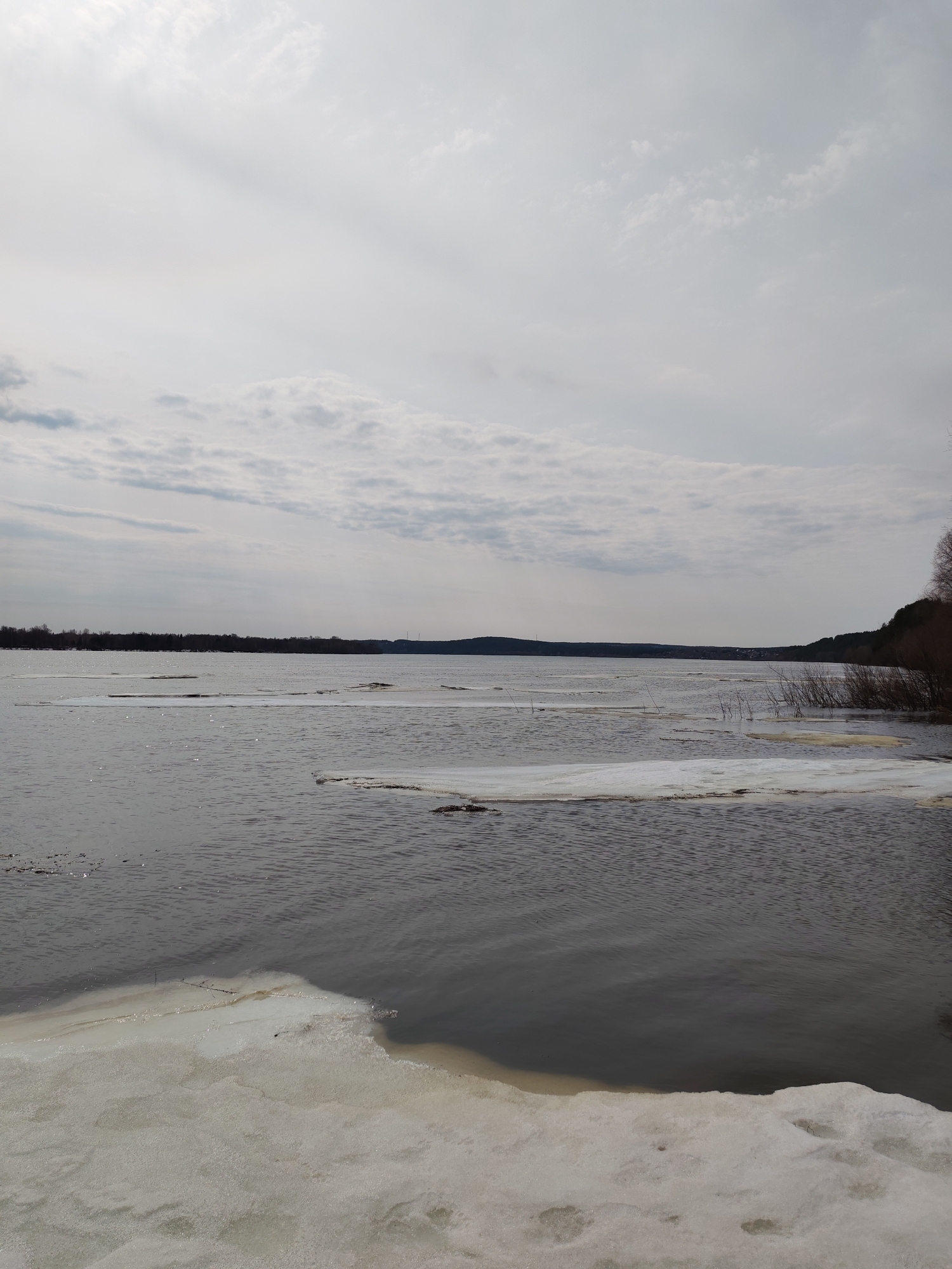 The image shows a partially frozen body of water, likely a lake or river, under an overcast sky. Patches of white ice are visible on the water surface and along the near shoreline. Bare trees and vegetation line the far shore and a section of the near shore to the right. In the background, the opposite bank appears wooded. The location is indicated to be in or near Поварёнки, Russia, based on the available information. The scene depicts the transition between winter and spring, with melting ice and dormant vegetation. No people are visible in the image.