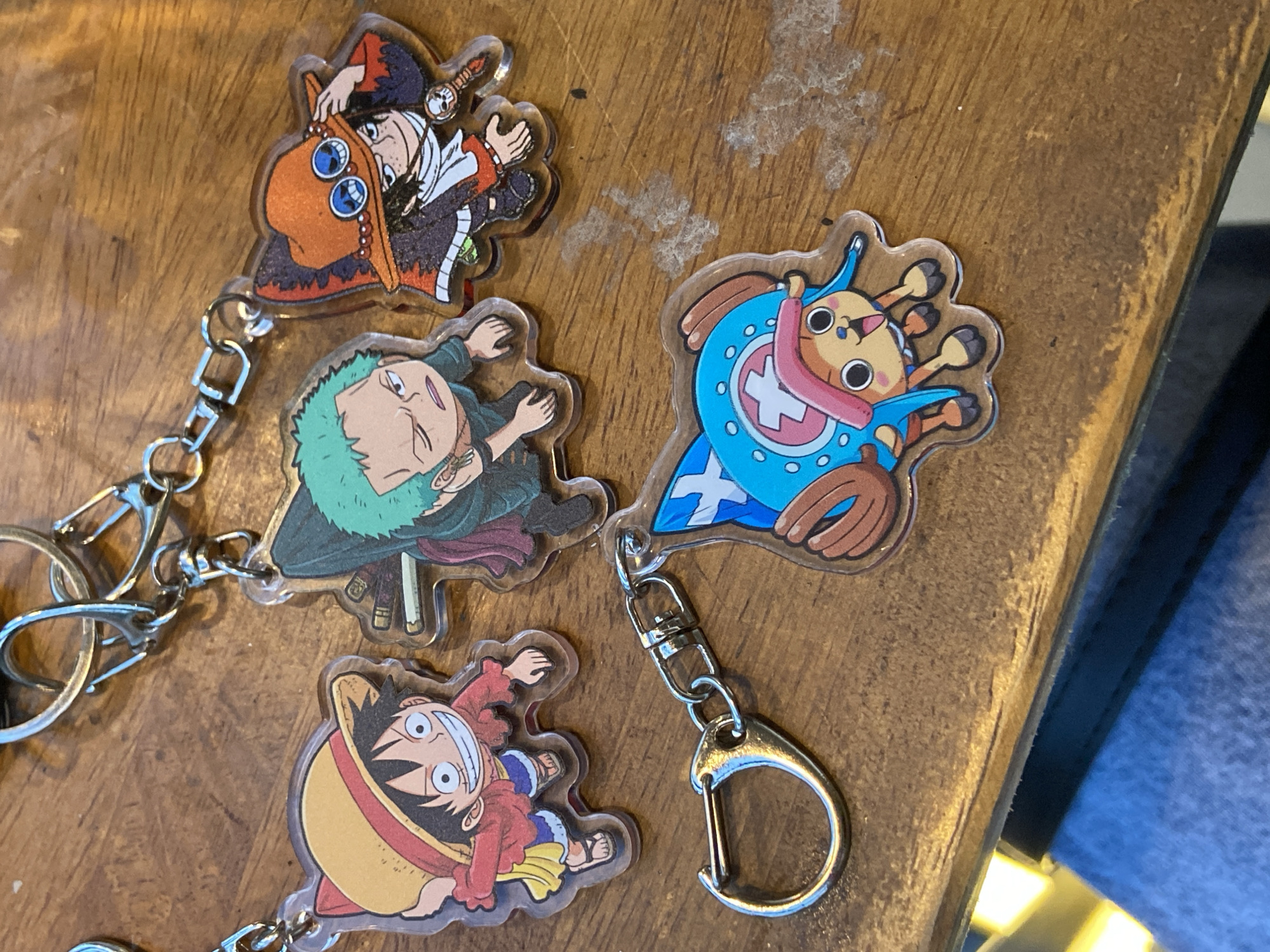 Four One Piece character keychains are displayed on a wooden surface. The keychains feature Monkey D. Luffy wearing his straw hat, Roronoa Zoro with green hair, Portgas D. Ace in his signature hat and outfit, and Tony Tony Chopper in his hat and blue coat. The keychains are made of clear acrylic and are attached to metal keyrings and clasps. The wooden surface appears to be a table with a visible grain and some dust or debris. The lighting suggests an indoor environment. This image does not provide any information that can be used to determine its location in Candirejo, Indonesia.
