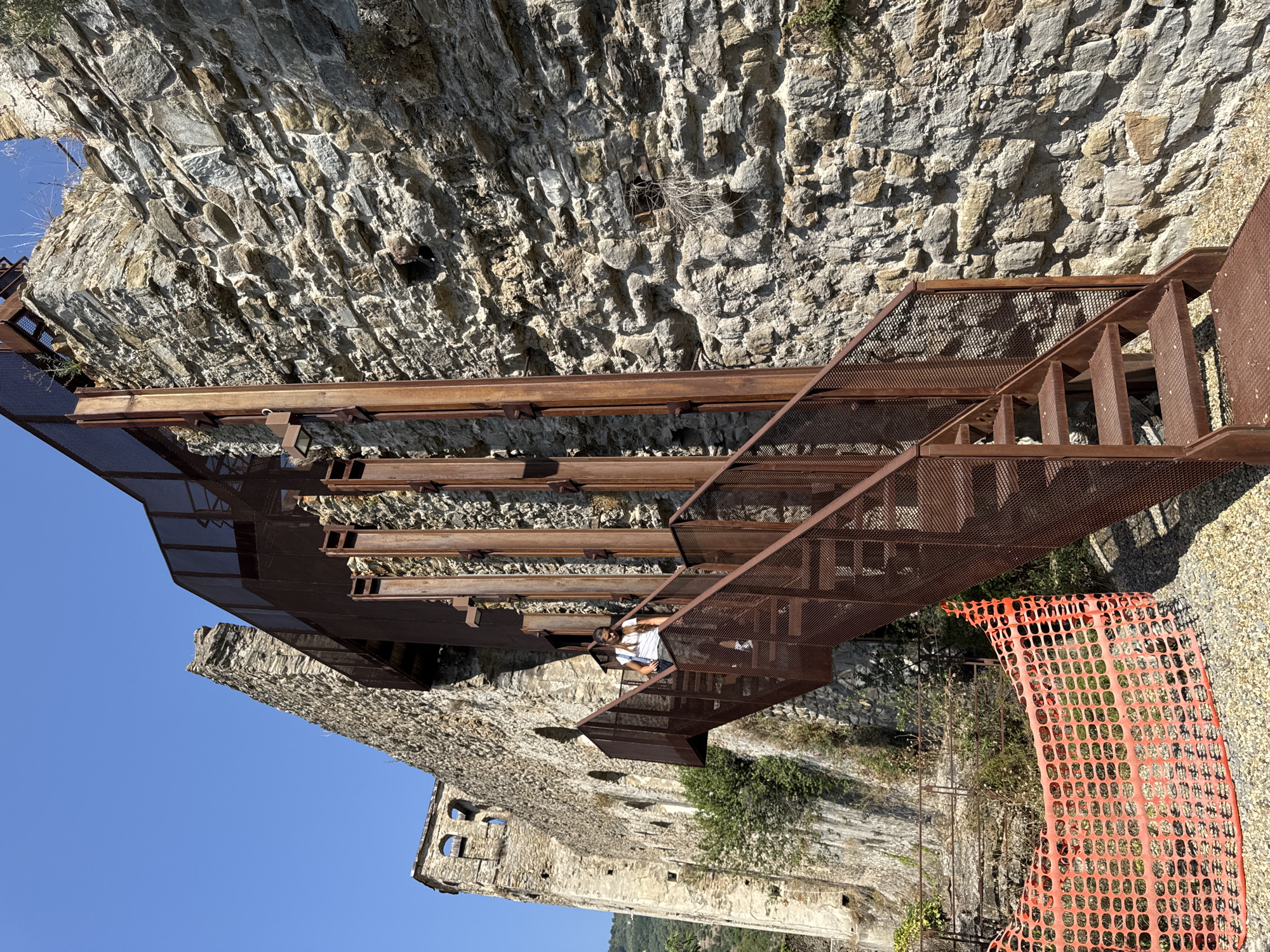 A woman walks down a rusty metal staircase attached to a stone building in Dolceacqua, Italy. The building appears to be old and possibly in disrepair, with parts of the stone wall crumbling and overgrown with vegetation. A bright orange plastic safety fence is visible in the foreground. The sky is clear and blue, indicating a sunny day. The staircase and its supporting structure are made of dark, weathered metal, with a grated surface for the steps and railings. The woman is wearing a white t-shirt and dark pants, carrying a backpack. The stone wall itself is constructed from irregular-shaped stones of varying sizes and colors.