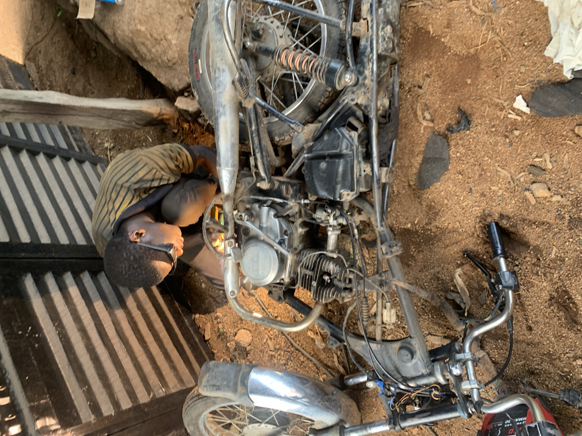 In Barakin Lerawi, Nigeria, a person wearing a striped shirt is working on a dirt bike. The scene is outdoors, with the ground covered in dry soil and loose stones. A large, rough-hewn rock formation is visible in the background, along with some wooden structures and a corrugated metal surface. The dirt bike is disassembled, with various parts scattered around it. The person is leaning over the bike, appearing to be focused on its repair. The lighting suggests it is daytime, likely morning or afternoon, with the sun casting shadows. The overall atmosphere is one of diligent work in a rural setting. No visible text is discernable in the image.
