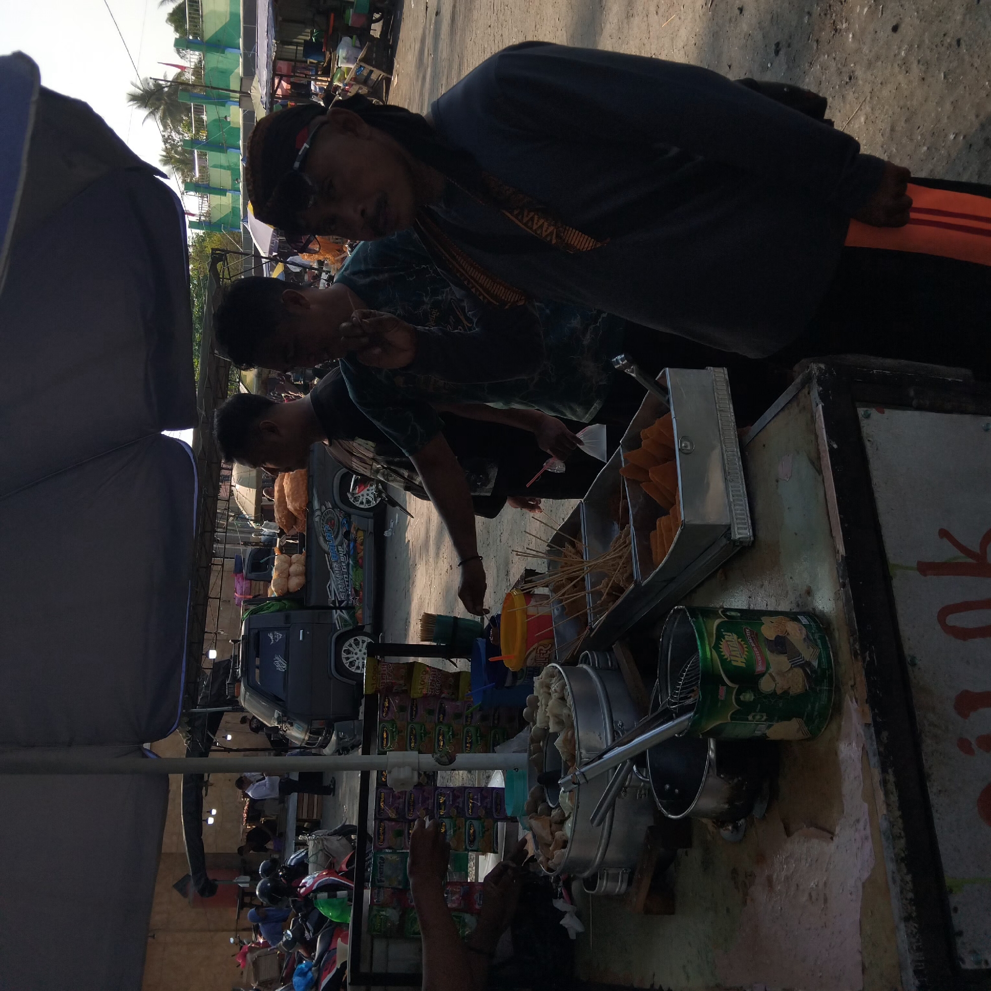 This is a street scene in Banjarbaru, Indonesia, likely a market or roadside food stall area. The foreground features a food cart with various items for sale. A large metal steamer basket is filled with meatballs and other food items, with tongs resting nearby. Next to it are metal trays containing skewers of fried food and what appear to be fried dough or snacks. A green tin can, labeled "HATARI ASSORTED BISCUITS", is also visible on the counter. Several people are present. Three men are standing near the food cart. One man, wearing a dark long-sleeved shirt and a patterned headband, is in the right foreground. To his left, another man in a dark t-shirt looks away from the camera. A third man, wearing a dark t-shirt with a graphic print, is reaching towards the food cart, possibly preparing an order. In the background, motorcycles are parked. A dark-colored pickup truck is also visible, adorned with a colorful decal on its side. Beyond the market area, there's a wall with a green and brown camouflage-like pattern and palm trees indicating a tropical or subtropical environment. The overall lighting suggests it is daytime, possibly late afternoon given the shadows.