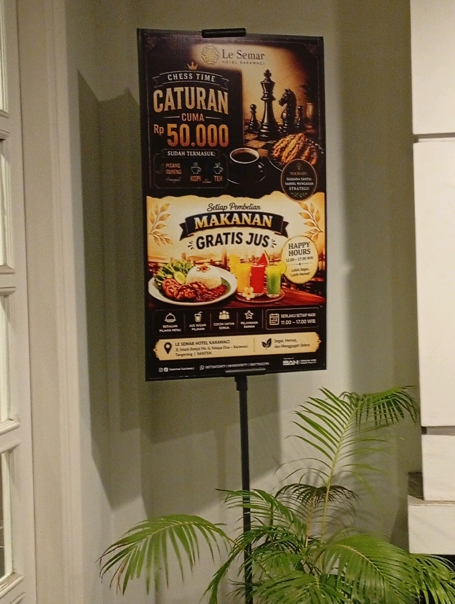 A promotional poster for "Le Semar Hotel Karawaci" is displayed on a black stand in front of a light-colored wall. The poster advertises a "Chess Time Caturan" offer for Rp 50,000, which includes food and a free juice. The food appears to be a rice dish with chicken and vegetables. The poster also mentions "Happy Hours" from 11:00 to 17:00 WIB. Below the main advertisement, contact information is provided, including the hotel's address: Jl. Imam Bonjol No. II, Kelapa Dua, Tangerang, Banten. A small palm plant is positioned in the lower right foreground of the image.