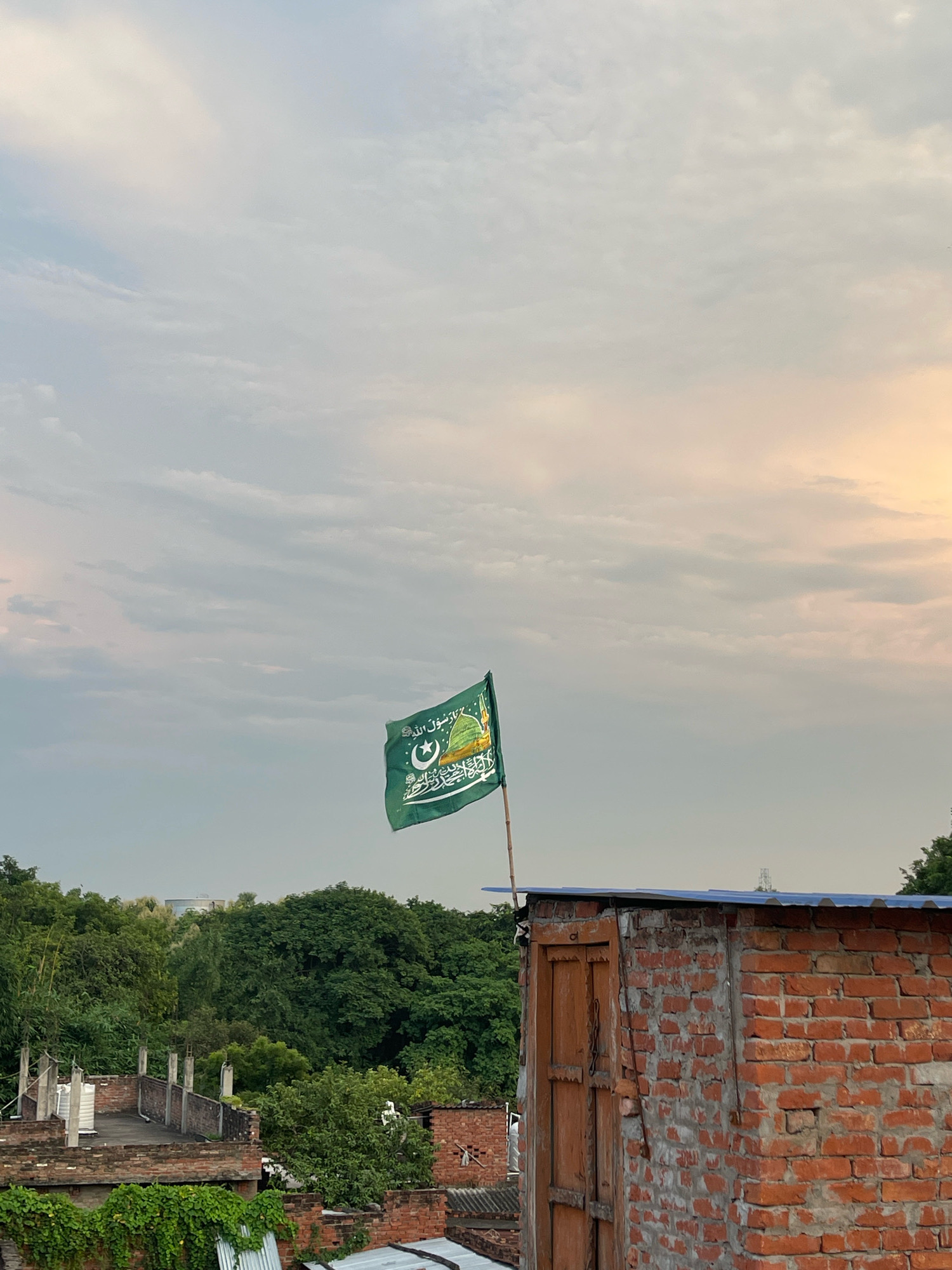 An Islamic flag, predominantly green with Arabic script and a crescent moon and star emblem, flutters from a bamboo pole atop a rustic brick structure. The structure features a wooden door and a corrugated metal roof. In the background, lush green trees form a dense canopy, with glimpses of other rooftops and structures in a city setting. The sky above is filled with soft, diffused clouds, with a hint of warm sunlight breaking through on the right side, suggesting either dawn or dusk. The image appears to be taken in Bojhi, India, based on the provided context.
