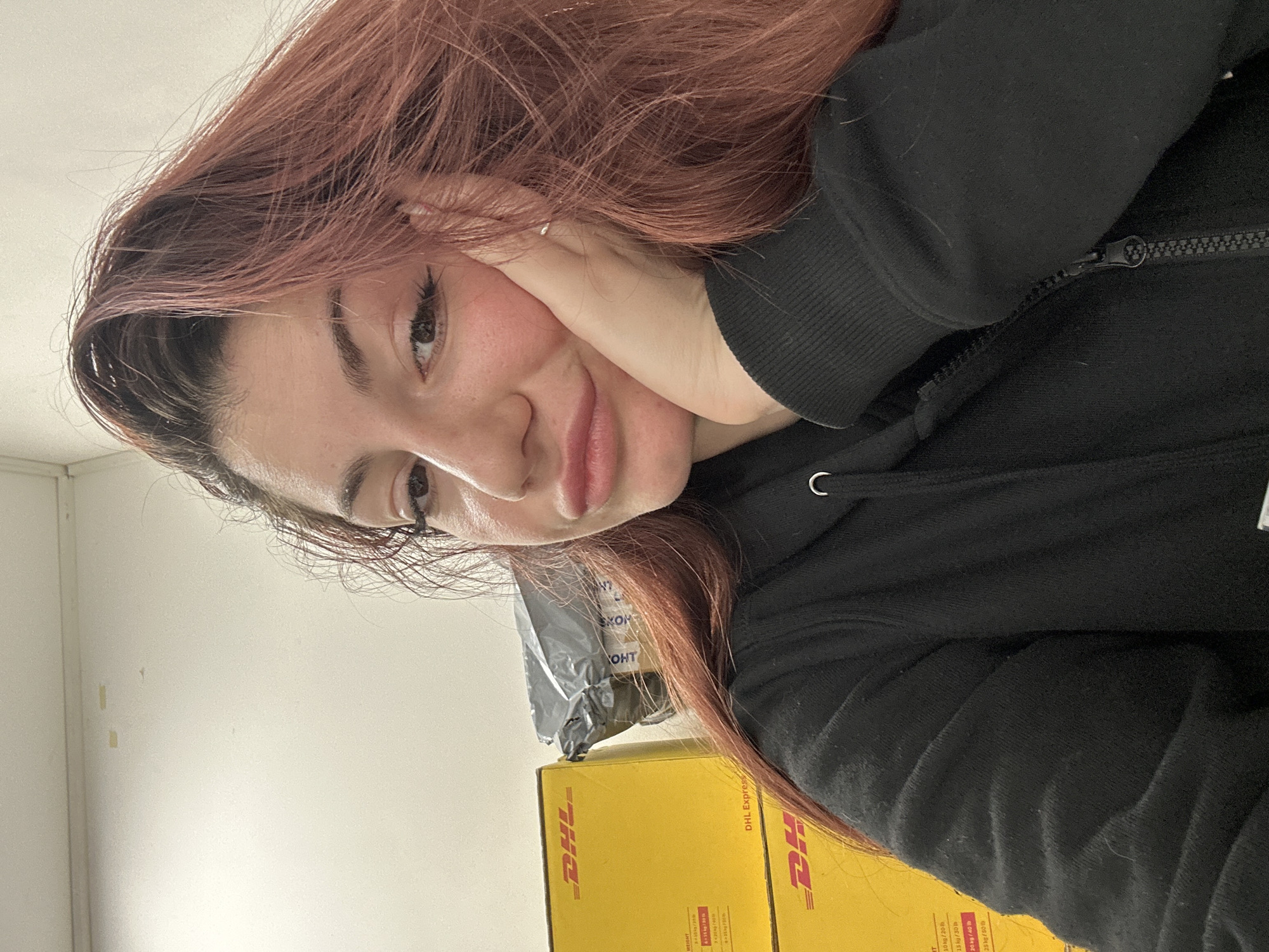 A young woman with reddish-brown hair rests her face on her hand, posing for a selfie. She wears a black hooded sweatshirt, zipped up. Behind her are two yellow DHL Express boxes and a gray package. The background is a plain off-white wall. There is no information in the image to identify the location as Varna, Bulgaria.