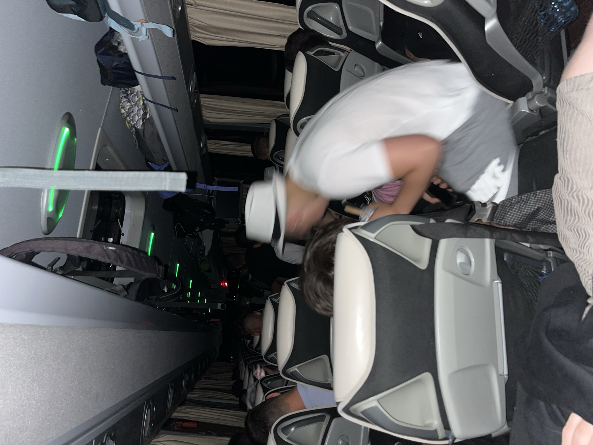 The image shows the interior of a bus, possibly in transit. Passengers are seated in rows of black and grey seats, with overhead luggage compartments visible. A person wearing a white hat and white shirt is bending over, appearing to reach into a bag or under the seat. The location is not discernable in the photograph, and therefore the relevance to Popovitsa, Bulgaria cannot be confirmed.