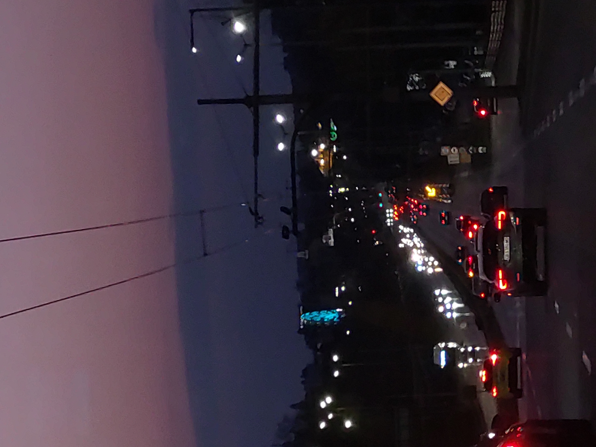 The image shows a street in Sofia, Bulgaria at night. The sky is dark with a hint of purple. There are power lines and poles on the left side of the image. On the right, there is a road with many cars, most of which have their headlights and taillights on. There are streetlights and some buildings with lights visible in the background. There is a traffic sign visible on the side of the road, indicating traffic direction or a hazard. The scene depicts heavy traffic on the road.