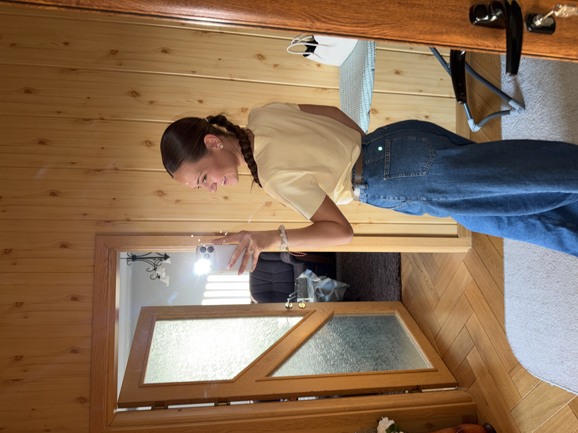 A female, appearing to be in her 20s or 30s, with dark hair styled in a braid, stands smiling and facing right, taking a mirror selfie with a silver smartphone held in her right hand. She wears a light yellow t-shirt, high-waisted blue jeans with a brown belt, and small white earrings. A light-colored beaded bracelet is on her right wrist. She stands on a light gray rug, on a wooden floor laid in a herringbone pattern. The walls in the immediate foreground are paneled with light-colored wood featuring prominent knots.
To the woman's right, a white handbag with a black interior rests on a light blue patterned surface, possibly an ironing board or narrow shelf, against the wooden wall. A dark wooden door frame with a black handle and a key is visible on the far right.
Reflected in the mirror, an open wooden door with frosted glass panels leads into an adjacent room. Through this doorway, a dark upholstered armchair is visible. A bright light source, likely a window with vertical blinds, casts a strong glare and streaks of light. A dark metal chandelier with three white shades hangs reflected in the upper portion of the mirror, above the doorway. The floor in the adjacent room appears to be dark gray carpet. No specific external markers confirming a precise location within Buhovo, Bulgaria are visible in this indoor scene.