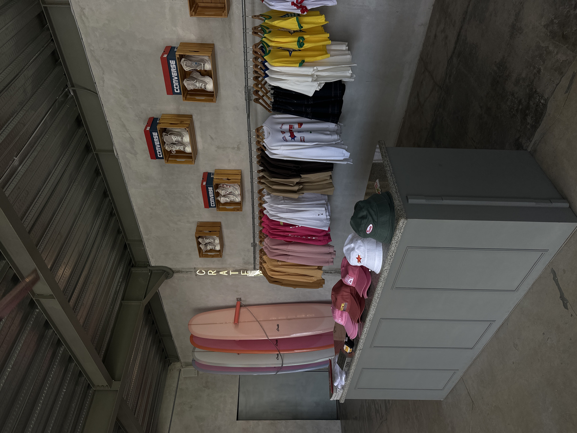 The image depicts the interior of a store in Mengwi, Indonesia, specializing in surf-inspired apparel and accessories. The store features a minimalist aesthetic with concrete walls and a corrugated metal ceiling supported by exposed steel beams. In the background, several surfboards in pastel colors are displayed vertically, leaning against the wall. Above a clothing rack, wooden crates are mounted on the wall, each showcasing a pair of Converse sneakers in their original boxes. The clothing rack displays a variety of casual wear, including hoodies, t-shirts, and long-sleeved shirts in a range of colors such as pink, yellow, white, and brown. In the foreground, a grey counter displays several caps, including pink and white styles, and a dark green bucket hat. The floor is made of concrete. No people are visible in the image.