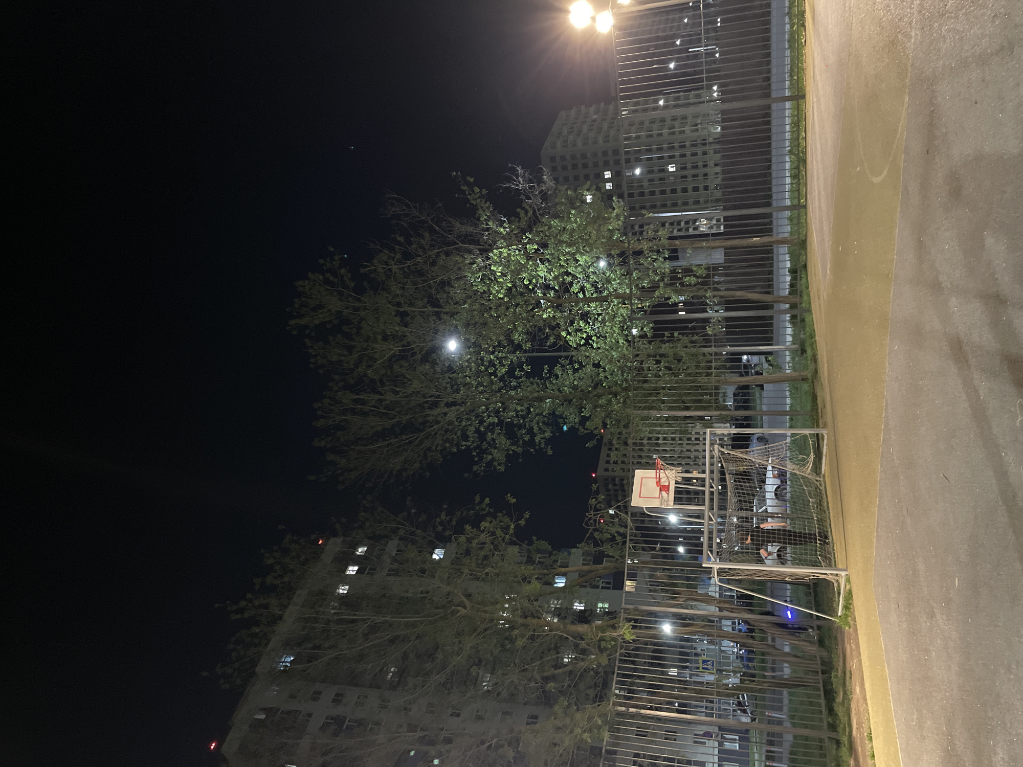The image shows a nighttime view of an outdoor basketball court located in Kazan, Russia. A metal fence surrounds the court, with trees and buildings visible in the background. The basketball hoop is visible on the side of the court and a soccer goal is placed on the court. Some light illuminates the area.