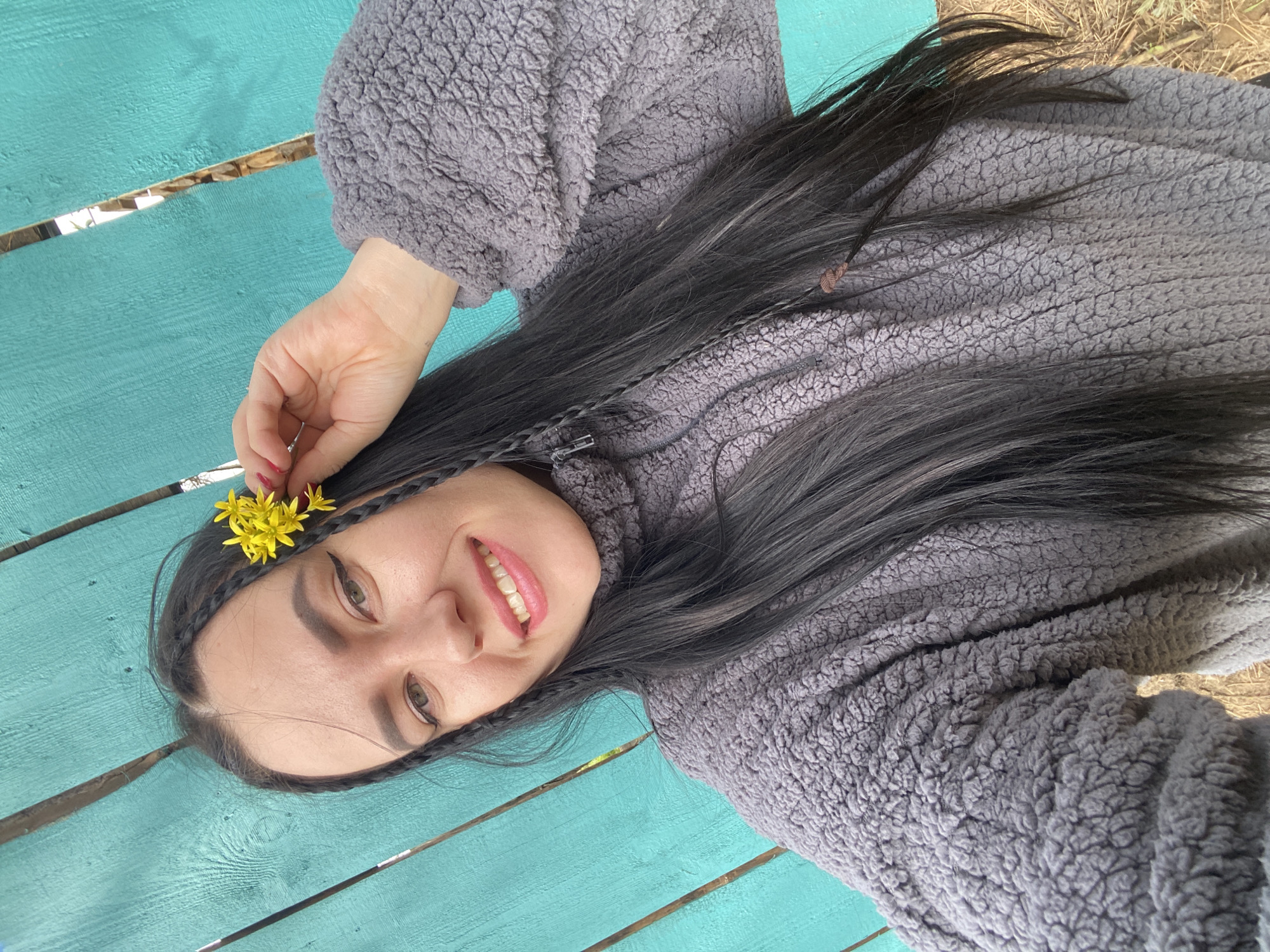 A woman with long, black hair adorned with braids and a small cluster of yellow flowers is lying on a turquoise-painted wooden surface. She is wearing a gray, textured fleece jacket or sweater. The location appears to be outdoors. The city is Chaikovsky, Russia.