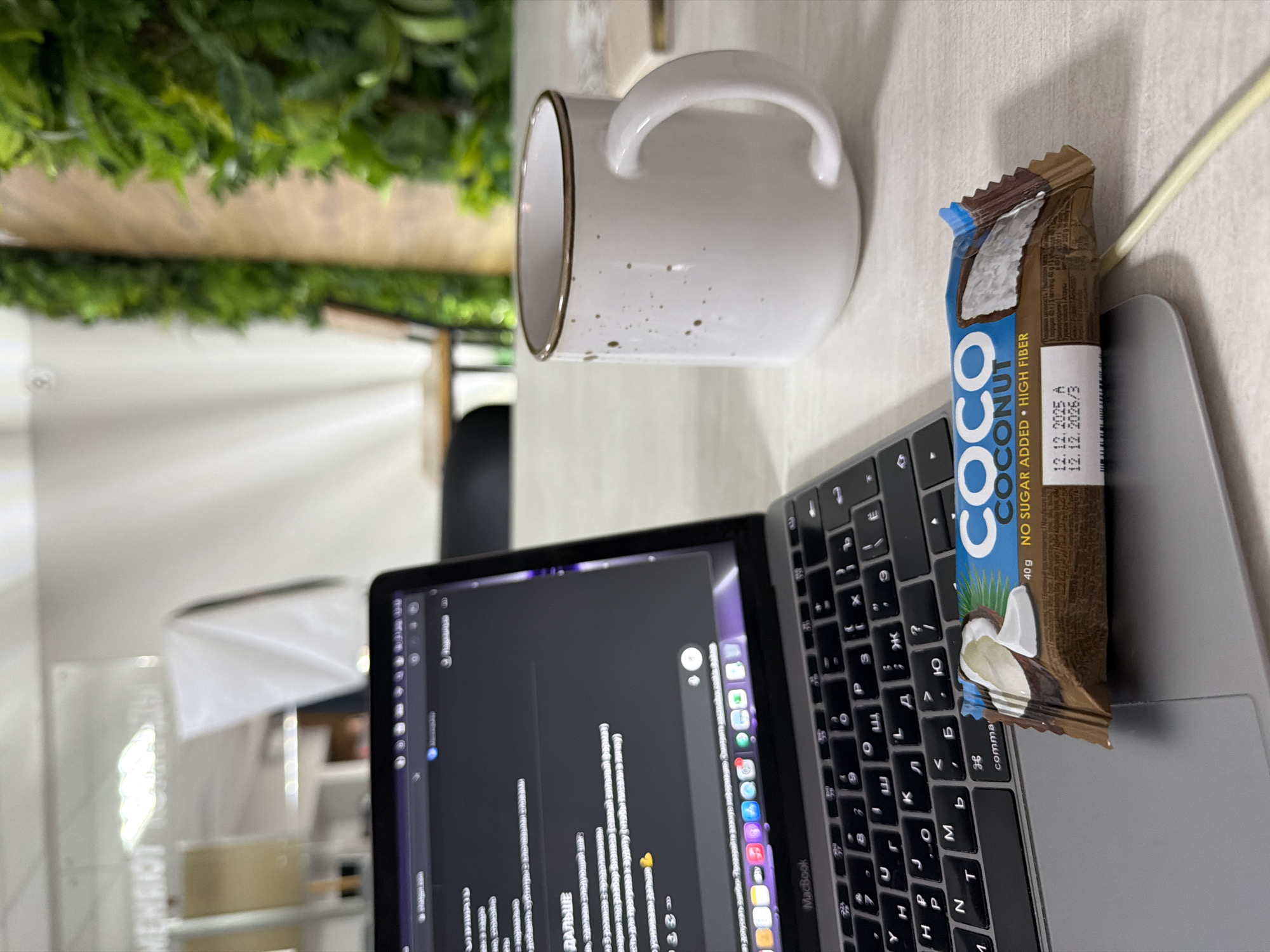 An overhead shot captures a detailed view of a workspace, likely an office or home setting, with objects arranged on a light-colored desk. The central item is a silver MacBook laptop featuring a dark grey keyboard. The keyboard displays both Latin and Cyrillic characters, indicating its use in a Russian-speaking region. The laptop screen shows lines of code or text on a dark background, with visible text including "ДАЛЬШЕ" (Further) and "от допускать ошиби" (from making mistakes), alongside the "MacBook" brand.
Resting partially open on the laptop's keyboard is a "COCO COCONUT" snack bar. The packaging highlights "40g NO SUGAR ADDED HIGH FIBER" and displays a manufacture date of "12.12.2025 A" and an expiry date of "12.12.2026/3". To the left of the laptop, a white ceramic mug with a brown rim and subtle brown speckles sits on the desk. A light-colored cable runs from the right side of the frame, passing by the snack bar.
The background is softly blurred, revealing green foliage, possibly artificial plants, adorning a wall or partition, which adds a natural element to the indoor scene. Further back, blurry white shapes and a faint, reflective clear sign with indistinct text, possibly "НЕИБИЛОТ", are visible. The scene suggests a person is engaged in work or study, taking a break with a snack and a drink. The Cyrillic keyboard strongly aligns with the provided location context of Chaikovsky, Russia.