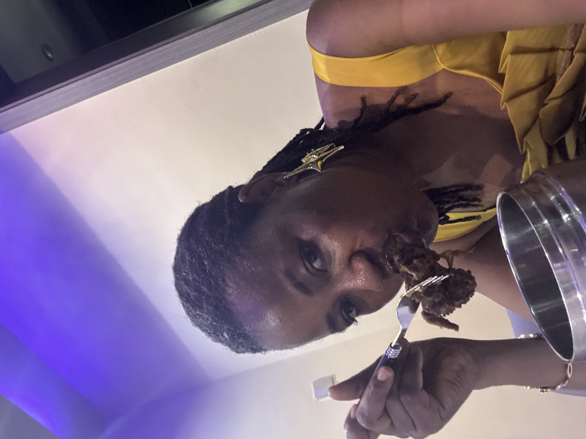 A woman with locs wearing a yellow top and star-shaped earrings is eating food from a fork held in her right hand. A silver bowl is partially visible next to her right arm. The location is not discernible to be Ajara, Nigeria from the visual details of the image. The background appears to be a generic indoor setting with white and blue-tinted walls.