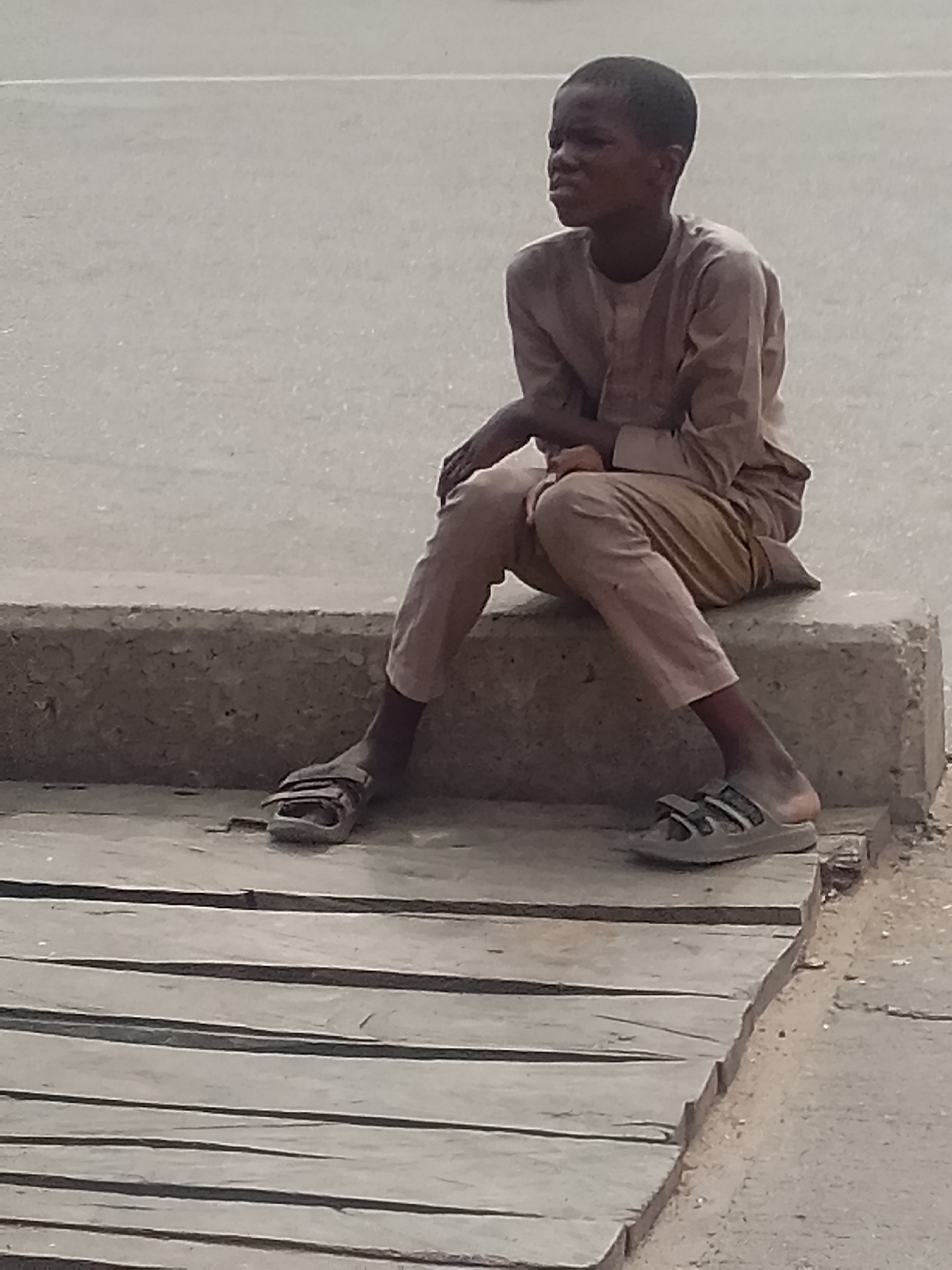 This image depicts a young male, appearing to be a boy or adolescent, seated outdoors in the city of Pantame, Nigeria. He is positioned on a light gray concrete ledge or low wall. The individual has dark skin and short, dark hair, and is dressed in a light brown, two-piece garment consisting of a long-sleeved tunic with a round neckline and matching trousers. Both feet are clad in light-colored, open-toed sandals. His body is oriented slightly to the right, and his head is turned towards the viewer's left, with his mouth slightly agape. His arms are clasped or resting in front of him. In the immediate foreground, below the concrete ledge, is a wooden platform composed of multiple horizontal planks. The background presents a uniformly light gray, paved surface, consistent with a road or open area, featuring a faint, lighter horizontal line in the upper portion of the frame.