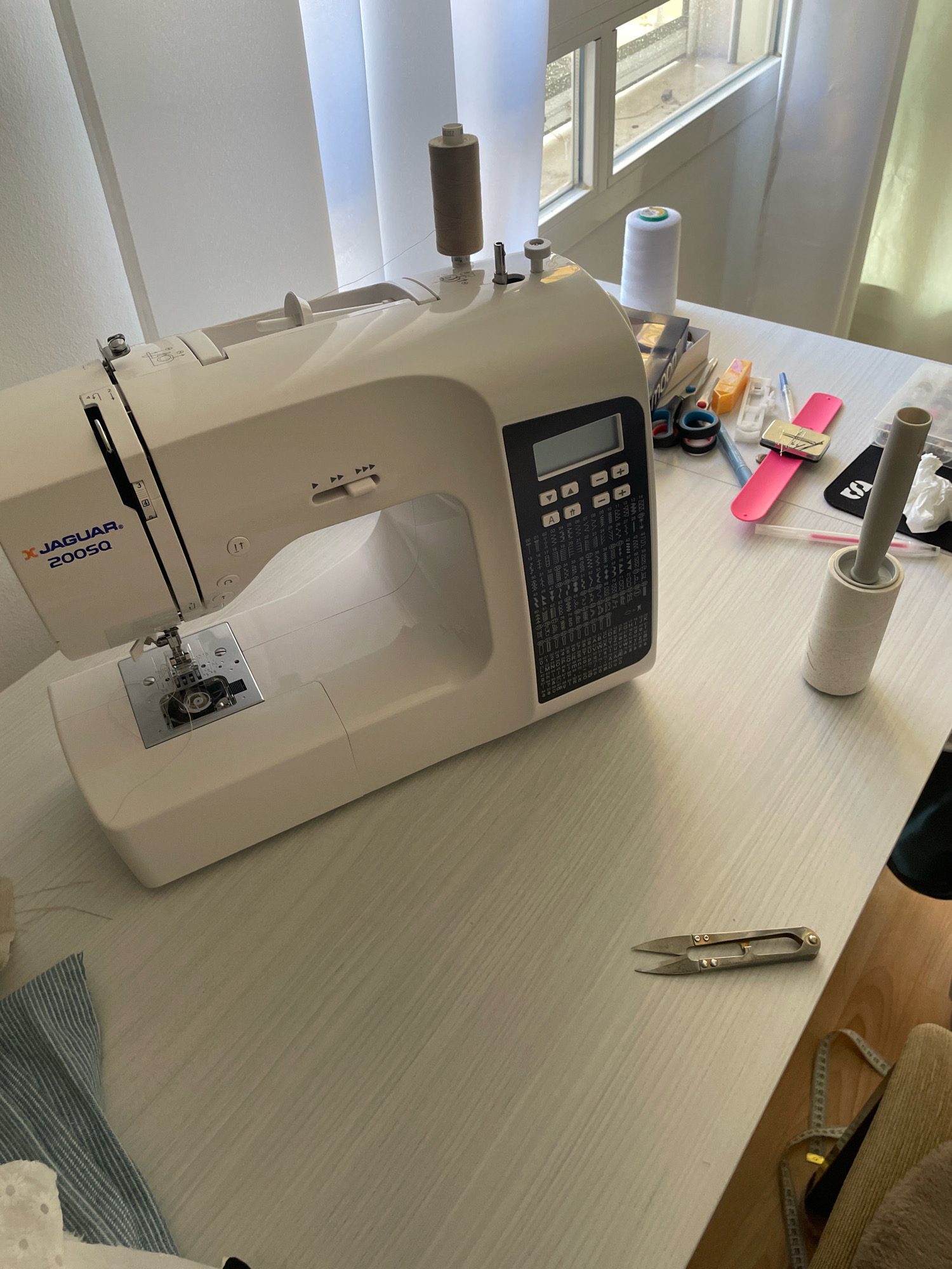 A white Jaguar 200SQ sewing machine is positioned on a light-colored, wood-grain textured desk. To the right of the sewing machine, a white spool of thread stands upright. A brown spool of thread is mounted on top of the sewing machine. Various sewing supplies are scattered on the desk, including a pink wristband with a magnetic pin holder, a pair of small silver scissors, a measuring tape, a lint roller, and a box of accessories. A patterned blue and white fabric is partially visible in the lower left corner. The background shows vertical blinds and a window with sunlight streaming through. The scene depicts a workspace for sewing.