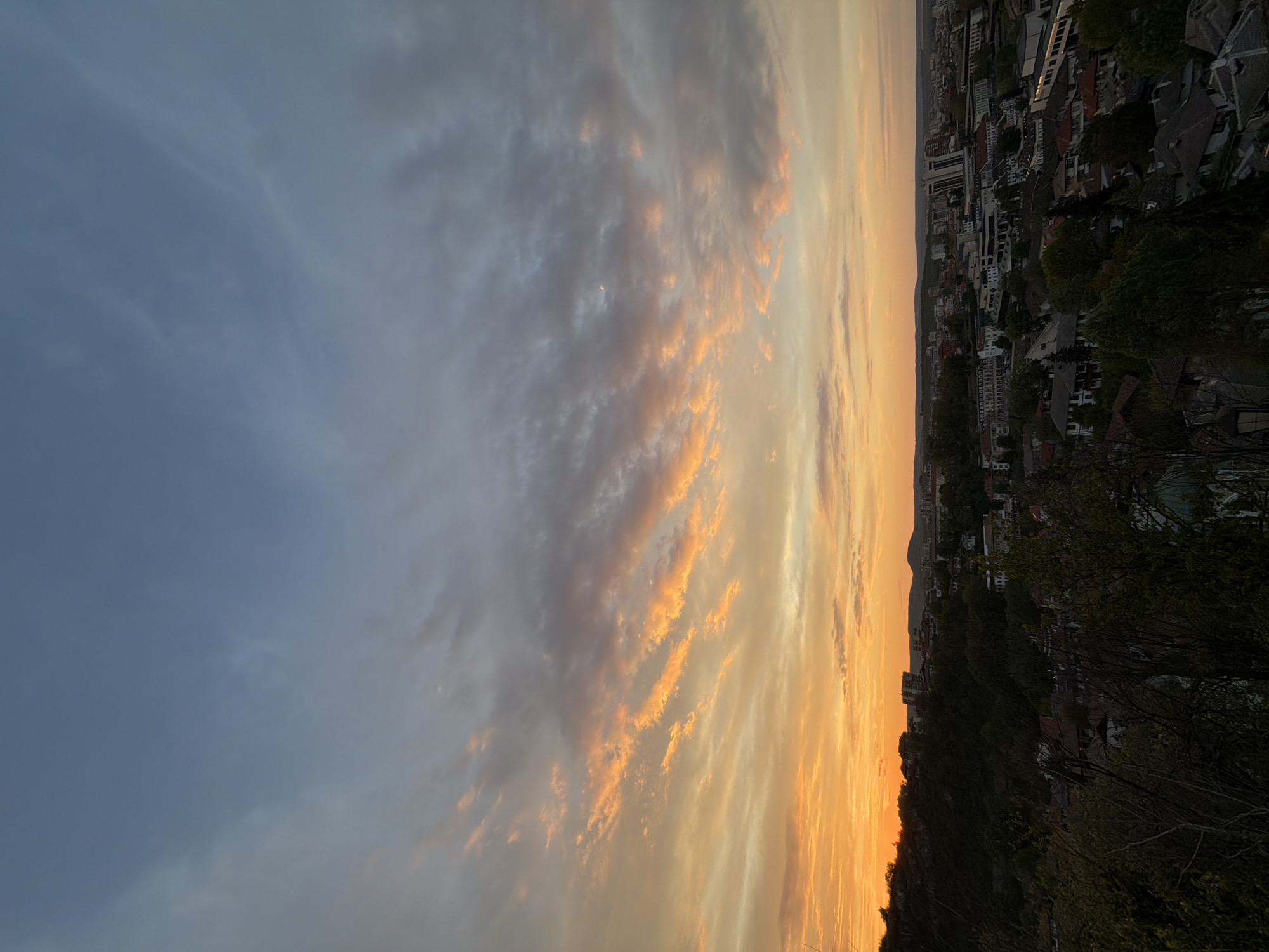 The image shows a cityscape, likely taken from a high vantage point, possibly in Lovech, Bulgaria. The sky is filled with clouds, transitioning from a blue hue to a warm orange and yellow near the horizon, indicating a sunset or sunrise. Buildings, trees, and a possible hillside fill the lower portion of the image, providing an urban/natural contrast. The image is rotated 90 degrees.