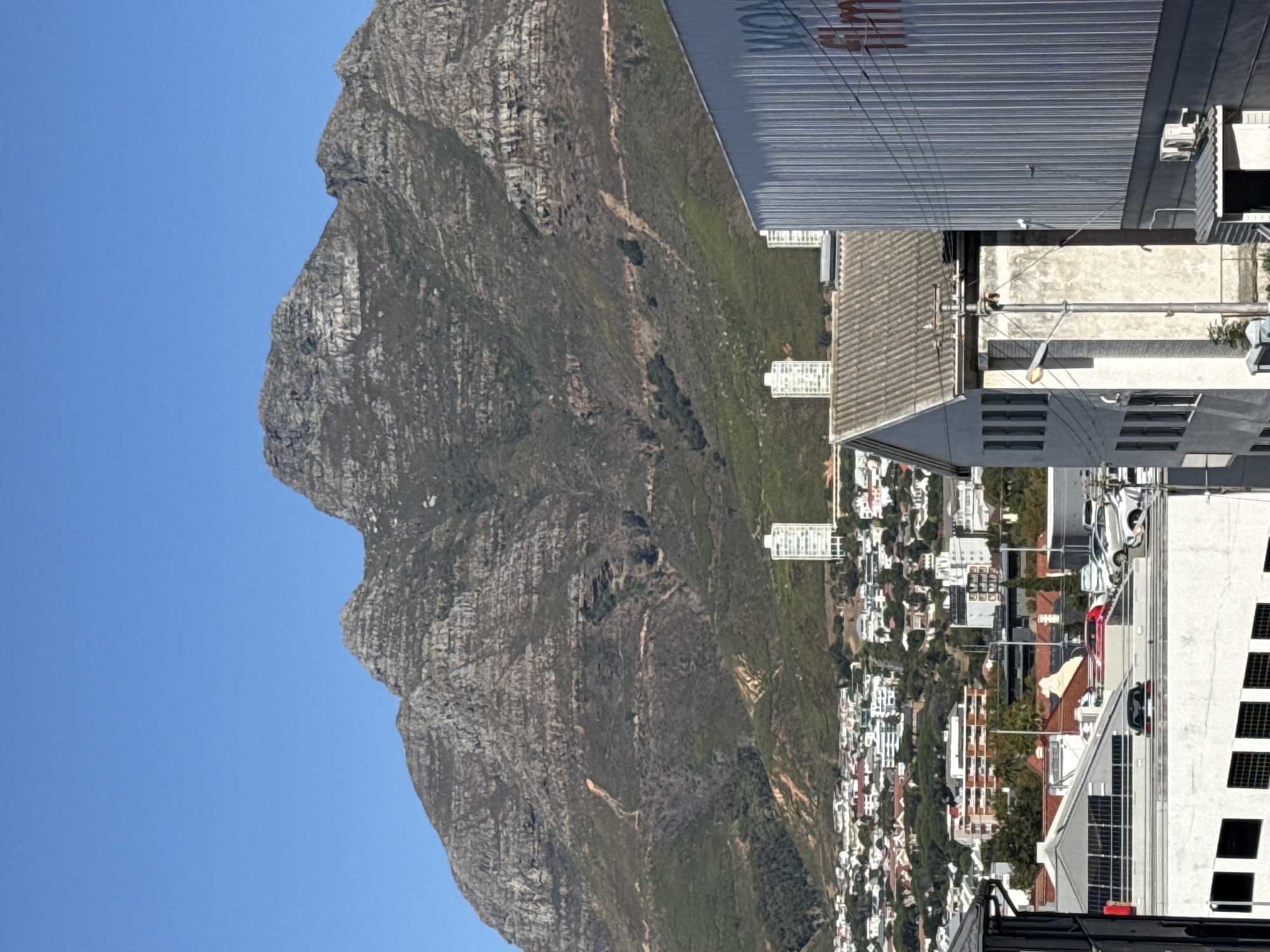 The image shows a landscape of Cape Town, South Africa, featuring Lion's Head mountain on the left side, set against a clear blue sky. To the right of the mountain, a densely populated area filled with buildings and houses stretches down the mountainside, indicating a suburban or urban residential zone. The foreground contains close-up views of the roofs of modern buildings, exhibiting various architectural elements such as solar panels and rooftop equipment.