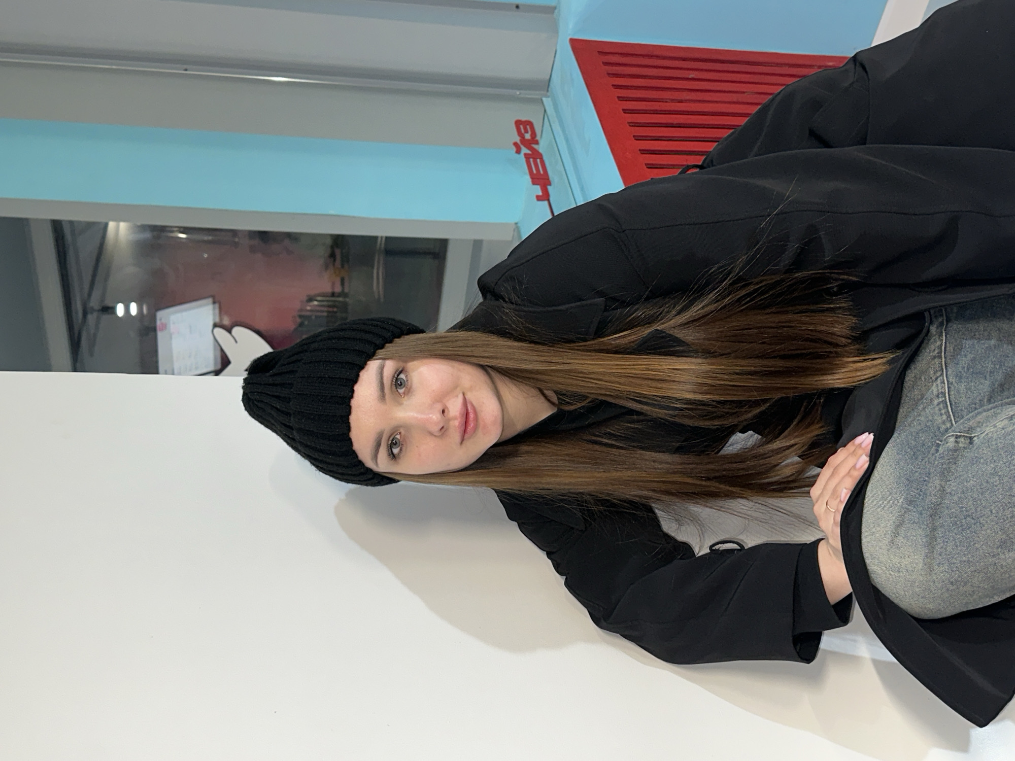 A young woman is depicted lying on a clean white surface, likely a floor or bench, gazing directly at the viewer with a soft smile. She has long, straight brown hair that cascades over her shoulders and is wearing a black ribbed beanie and a black long-sleeved jacket. Underneath her jacket, blue jeans are visible. Her hands are in view, with light pink nail polish on her fingernails and a simple ring on one of her index fingers.
The background features a light blue wall or partition. On the right side of the frame, a distinct red slatted structure, possibly a ventilation cover or decorative element, is mounted on the blue wall. Cyrillic text, reading "ЧЕЙЗ" (CHEYZ in English transliteration), is clearly visible on this red structure. To the left, a reflective surface, resembling a window or glass partition, shows a blurred reflection of what appears to be an indoor setting with a monitor or screen displaying bright points. The overall setting suggests a modern indoor environment, consistent with public or commercial spaces in Chaikovsky, Russia, given the visible text. The lighting is uniform, indicating an indoor artificial light source. The woman appears relaxed and approachable.