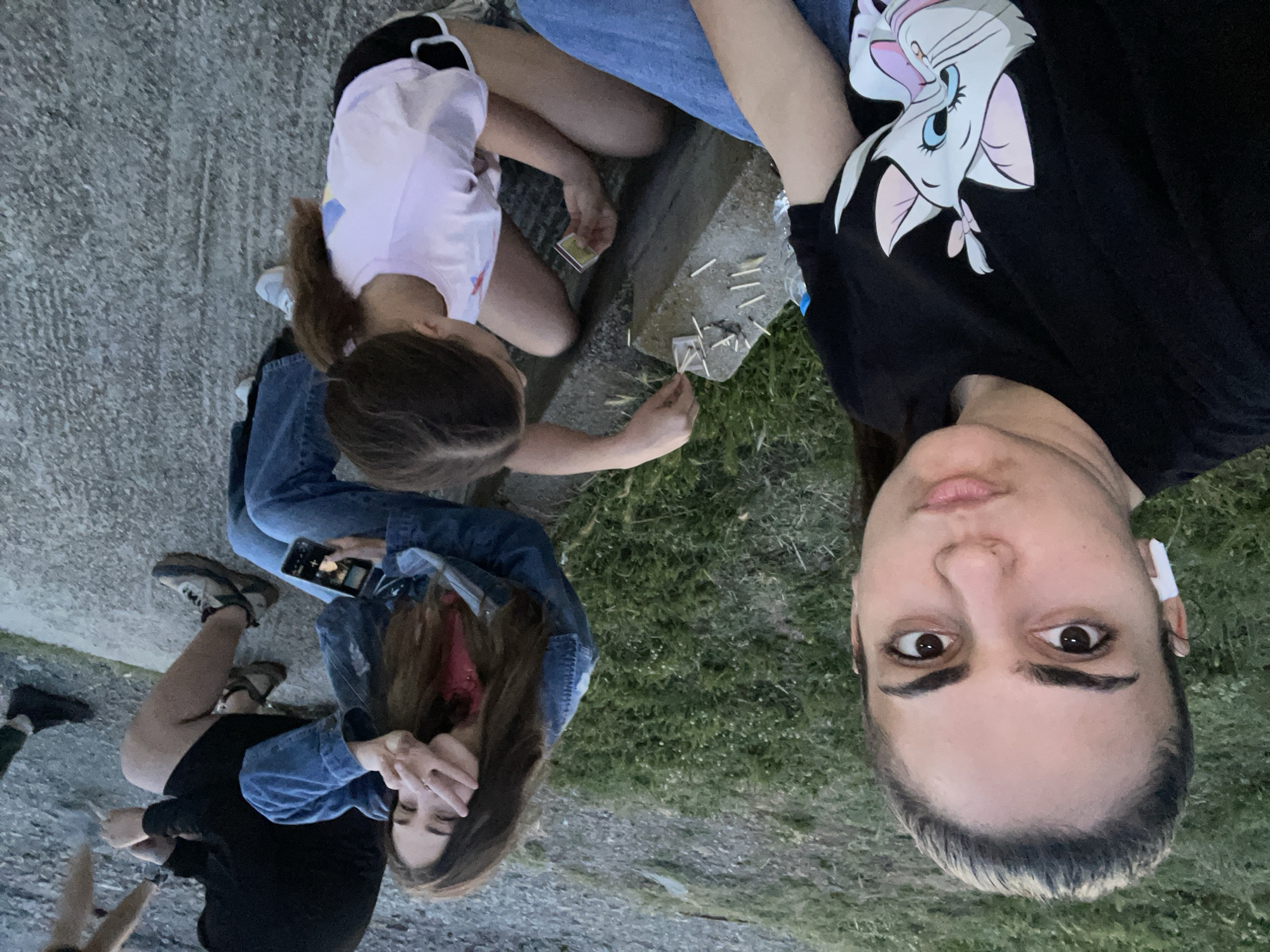 An outdoor scene in Debelt, Bulgaria, depicts five individuals gathered on and around concrete structures with grassy and gravel ground. In the foreground, a young woman with dark hair pulled back is taking a selfie, looking directly at the camera with a slight pout, wearing a black t-shirt with a "Marie" cat graphic and blue jeans, with a white wireless earbud in her left ear. To her right, a young girl with dark hair in a ponytail, wearing a light pink t-shirt and black shorts, is seated on a low step. She is holding an open matchbox in her left hand and arranging numerous small, light-colored wooden matchsticks on a grey concrete block positioned between her and the foreground woman. A plastic water bottle is partially visible behind the foreground woman's arm. Further back, a young woman with long dark hair, wearing a denim jacket over a pink top and blue jeans, is seated on a concrete ledge. She is holding a smartphone in her left hand, which displays an image on its screen, and making a peace sign with her right hand, partially obscuring her face. Behind her, a person wearing a black hoodie, black shorts, and athletic shoes is partially visible, seated on a similar concrete surface. Further to the right, only the legs and feet of two more individuals in dark clothing and shoes are visible. The ground shifts from green grass with small white flowers in the midground to a grey, gravelly or paved surface in the background. The ambient lighting suggests late afternoon or early evening.