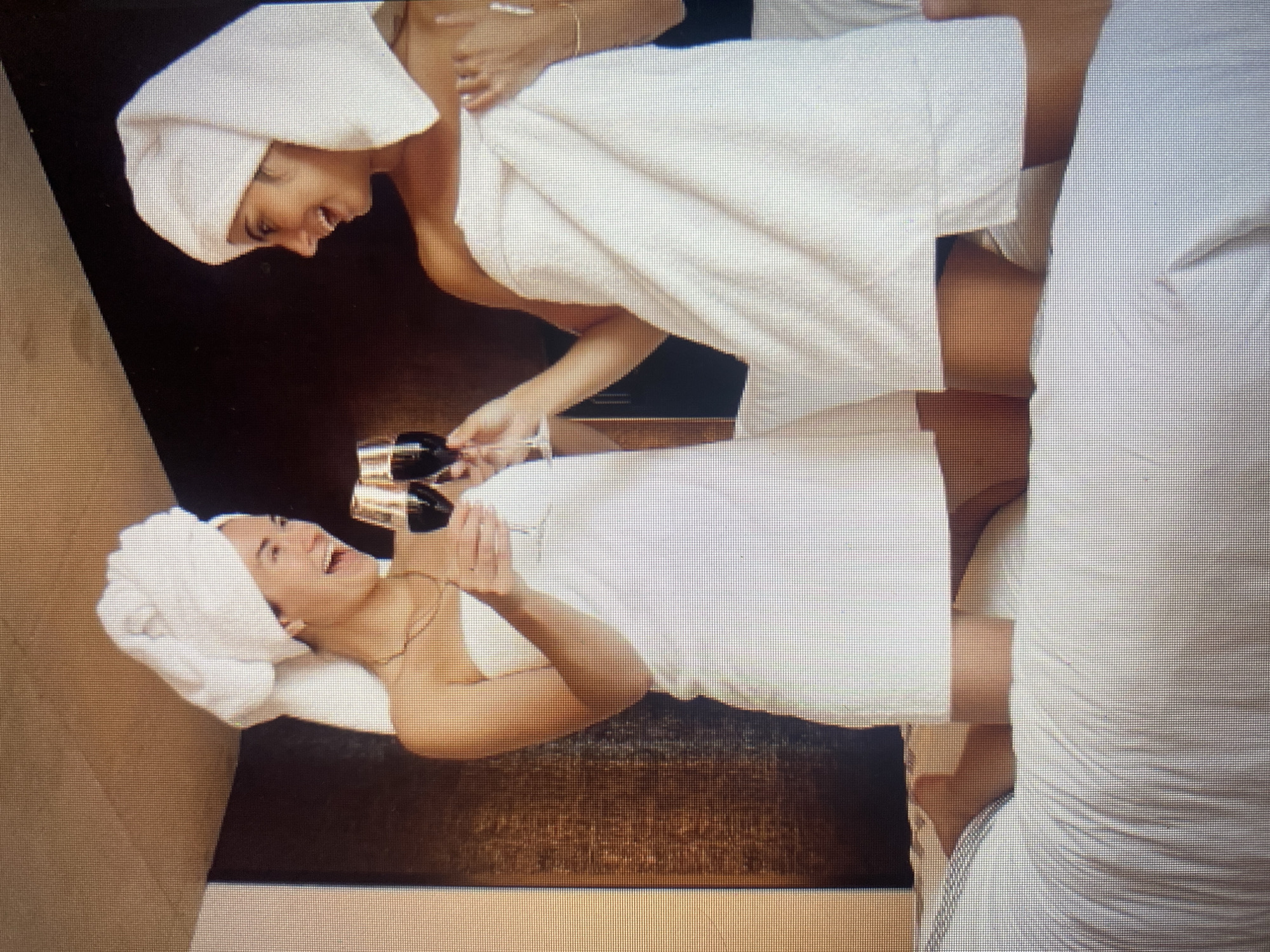 Two young women, wrapped in white towels and with towels on their heads, are seated on a bed. They are holding wine glasses and appear to be toasting. The room has a textured, wood-paneled wall in the background. The bed is covered with white linens.