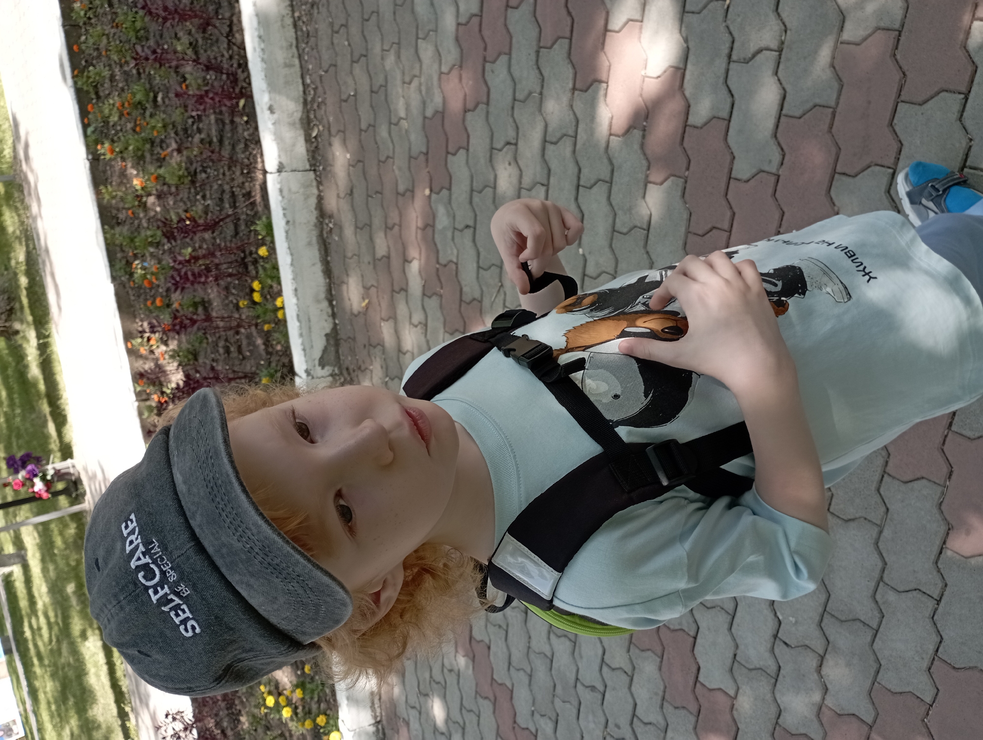 A young person with curly, light-colored hair, freckles, and brown eyes looks up at the camera. They are wearing a grey baseball cap with white text that reads "SELFCARE BE SPECIAL" and a light blue t-shirt with a graphic print. A black backpack with a green accent is visible on their shoulders.
The person is standing on a paved walkway made of light and dark interlocking stones. To the left of the walkway, there's a flower bed with small orange and yellow flowers, and some purple flowers can be seen in the background. The setting appears to be outdoors, possibly a park or a residential area, with shadows suggesting it's daytime.
The image is taken from a slightly low angle, looking up at the subject. The lighting suggests it could be a sunny day. The overall mood seems casual and relaxed, with the subject looking directly at the camera, perhaps posing for a photo. There is no specific event depicted, it appears to be a candid or posed portrait. The location context of Blagoveshchensk, Russia, provides geographical information but doesn't offer specific visual cues within the image itself.