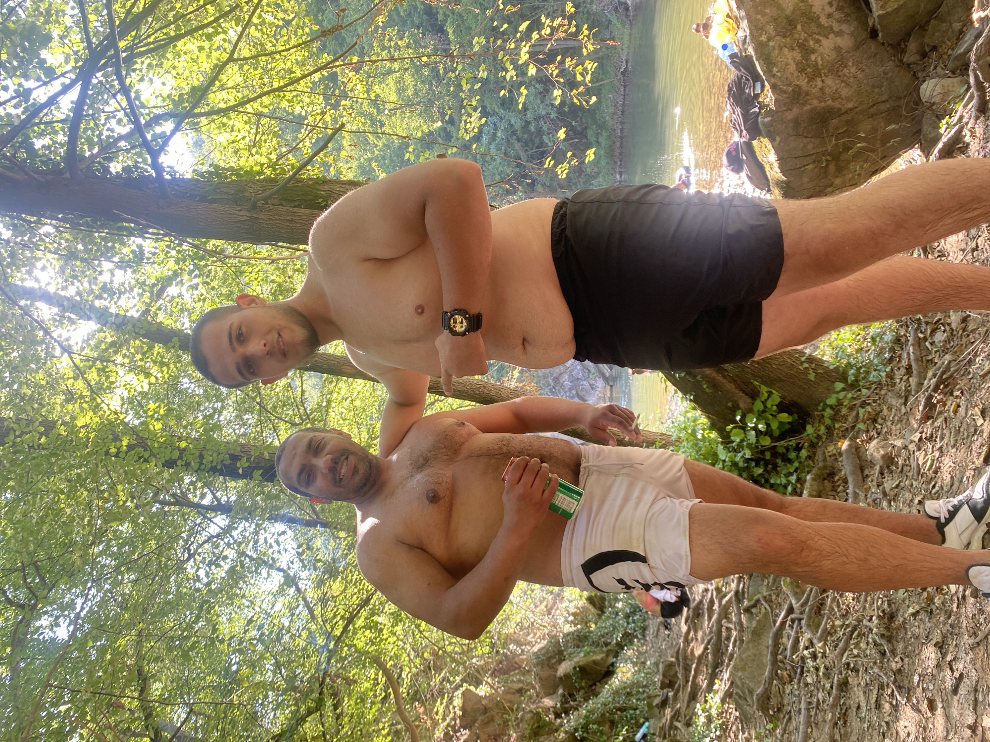 Two shirtless men stand near a river in Debnevo, Bulgaria. The man on the left, holding a green can, wears white shorts with black accents. The man on the right wears black shorts and a watch on his left wrist. The background features a river, trees, and sunlight filtering through the leaves.