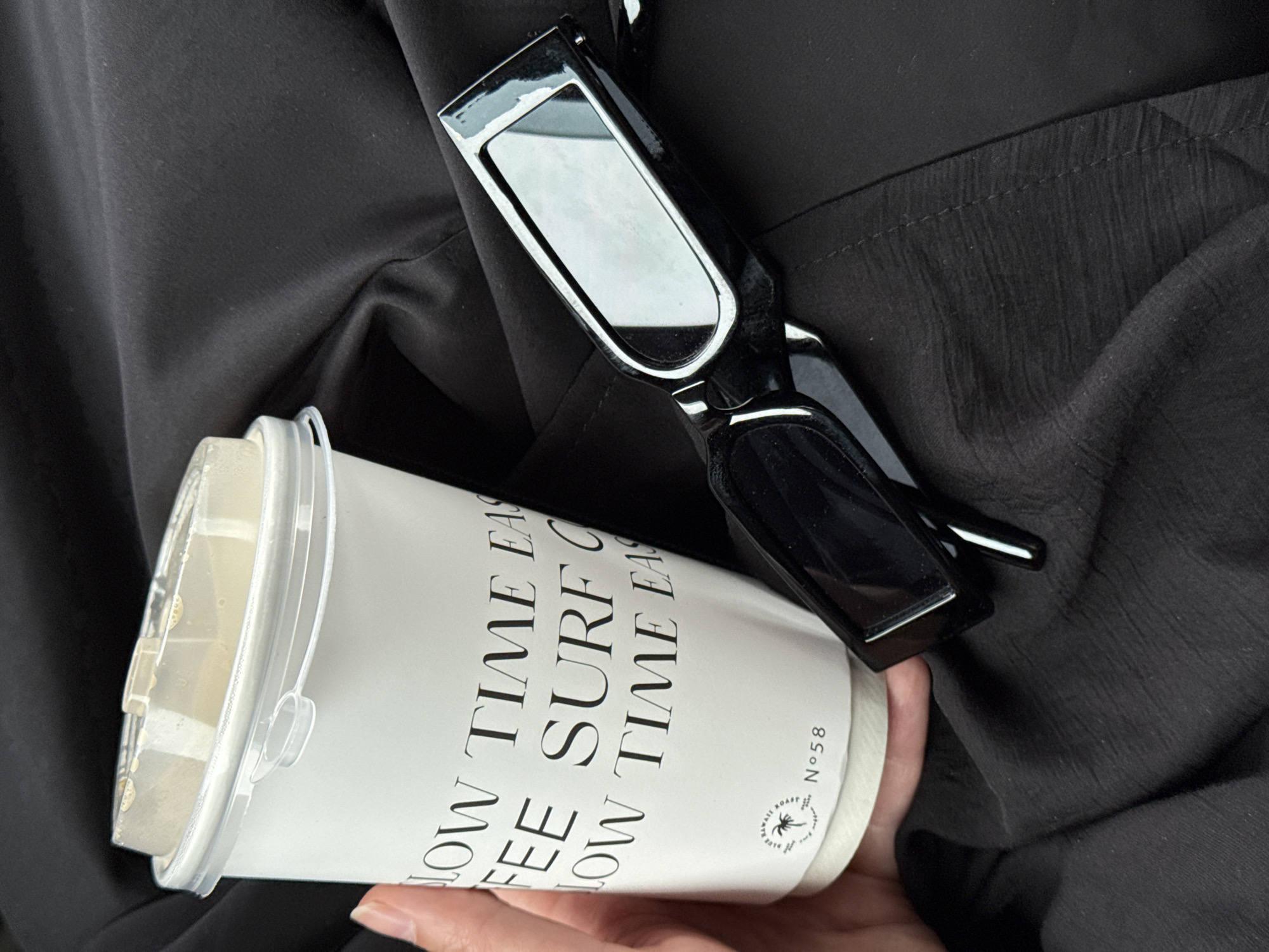 A photograph shows a person holding a white paper coffee cup with a clear plastic lid. Text on the cup reads "SLOW TIME EASY FEE SURF C LOW TIME EAS" and "Nº58." Below the text is a small circular logo with a palm tree graphic. A pair of black-framed sunglasses rests on the cup and black fabric. The person's fingers are visible beneath the cup. The background consists of draped black fabric. The image gives no information about Tavlarovo, Russia.