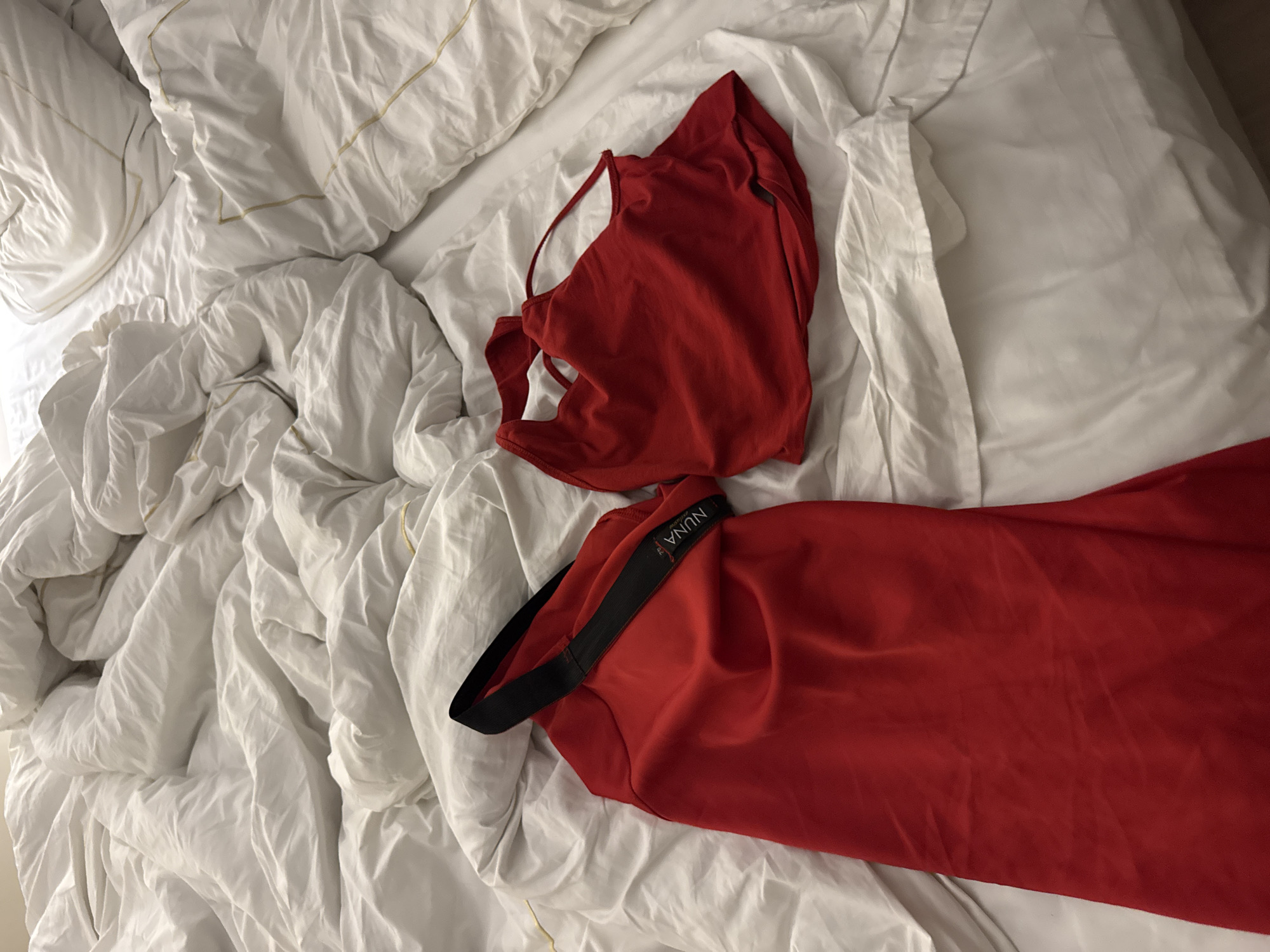 Two red garments are spread out on a rumpled white duvet. The garment on the left is a long, sleeveless dress with a black elastic band across the top. A label with the word "NUNA" is visible on the black band. The garment on the right is a red tank top. The scene is likely indoors, possibly in a bedroom, given the presence of the duvet. There is no indication of the location being in the city of Unknown, Egypt, or any specific city.