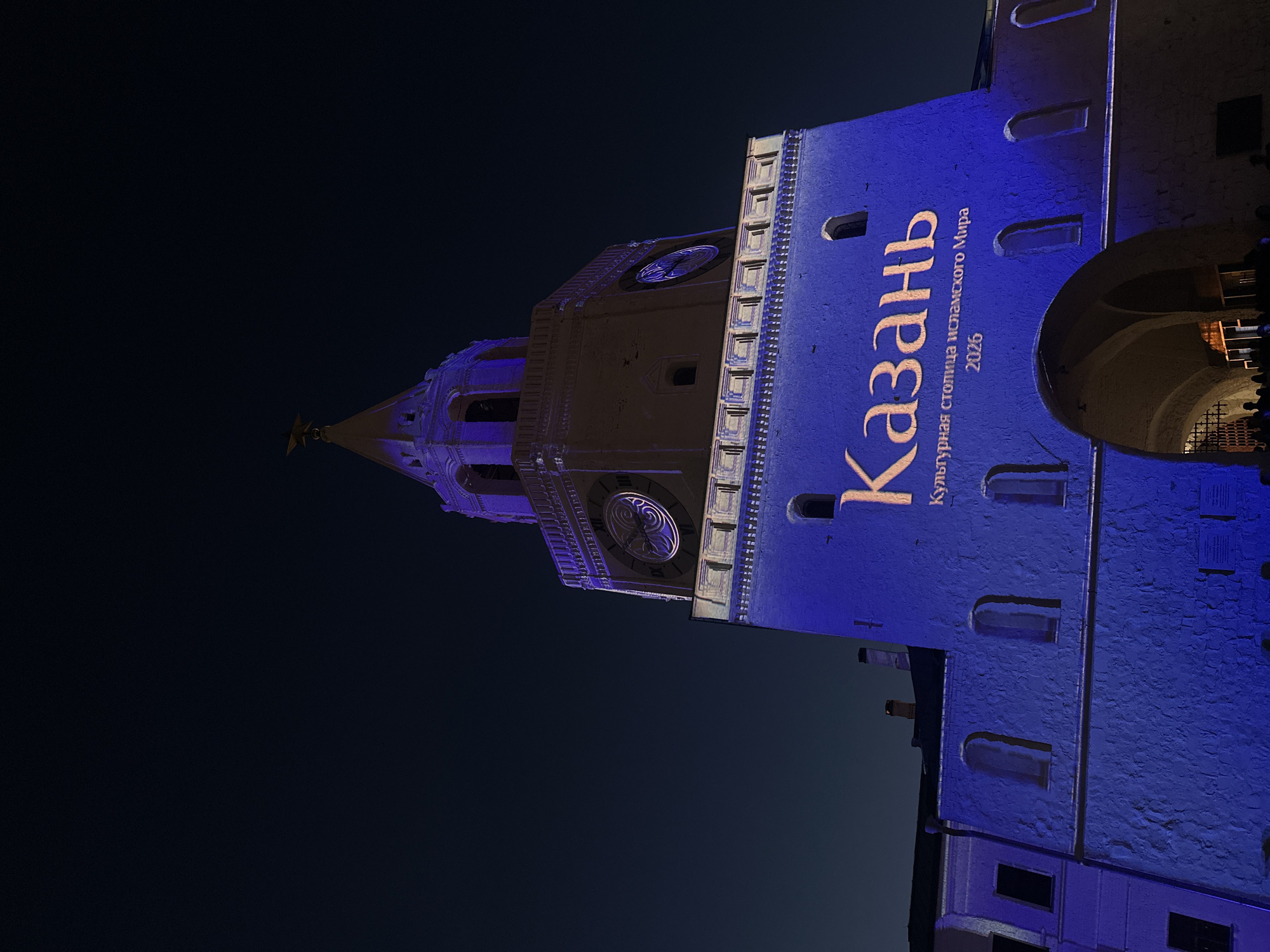 The image shows a building in Kazan, Russia, illuminated at night. A vertical inscription in Russian, "Казань культурная столица исламского мира 2026" (Kazan, cultural capital of the Islamic world 2026), is projected onto the building's facade in orange. The building itself is lit in blue. A clock tower is visible on the left side of the building, with its clock face illuminated. In the background, the sky is dark. A structure with an arched opening is visible on the right side of the building.