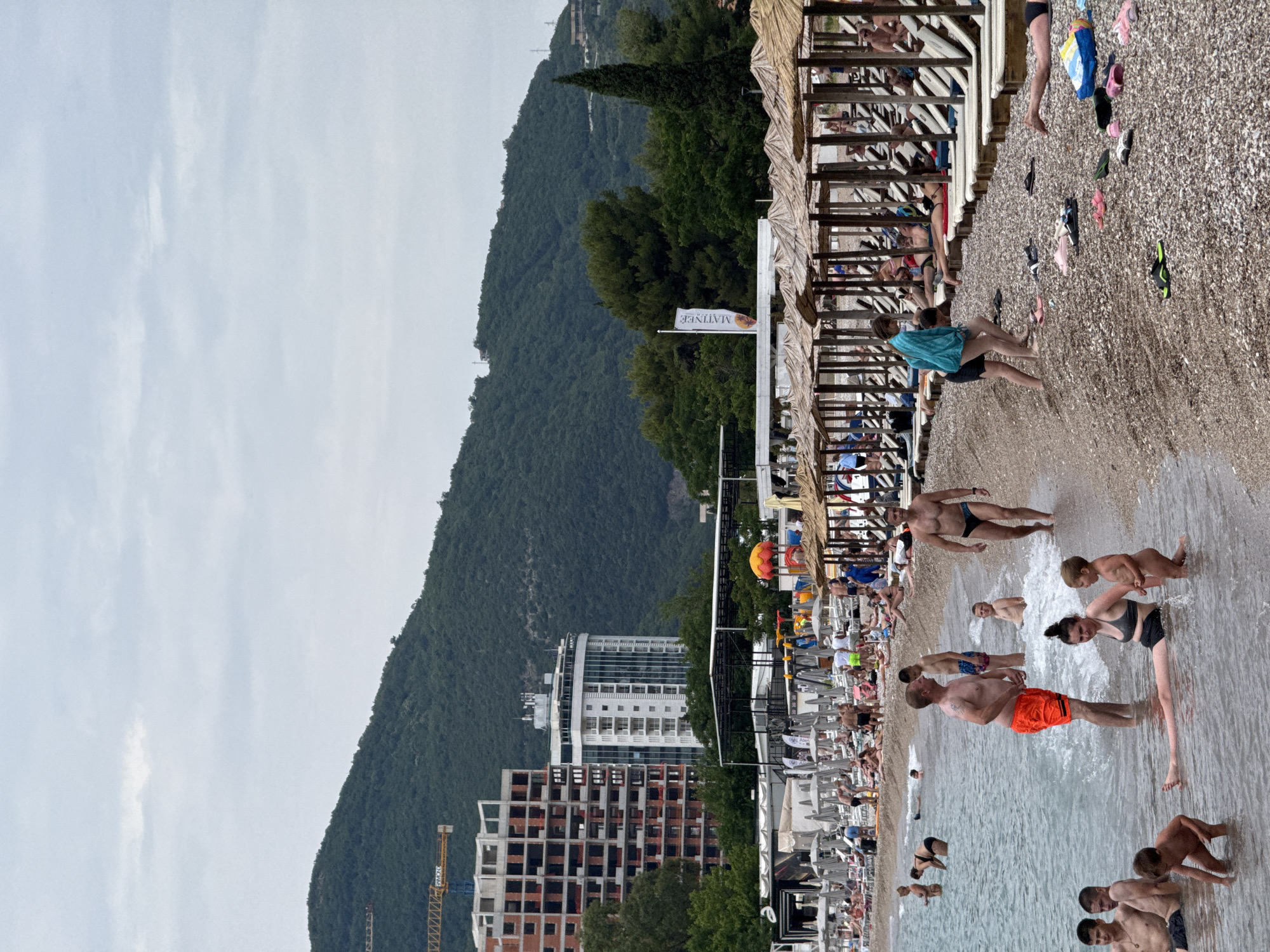 This image captures a vibrant daytime scene at a pebble beach in Budva, Montenegro, under an overcast sky. The foreground and mid-ground are teeming with people enjoying the sea and shore. To the right, individuals are wading and standing in the clear, shallow water of the sea, including a man in bright orange swimming shorts and others in dark swimwear. Children are also visible playing in the water.
Moving towards the left, the pebble beach extends, populated by more people either walking or relaxing. Rows of wooden sunshades with thatched roofs and sun loungers are set up along the beach, with many occupied. Various items of footwear, such as sandals and flip-flops, are scattered on the pebbles near the water's edge, alongside a blue and yellow beach bag.
In the background, lush green mountains covered in dense vegetation rise prominently, defining the natural landscape. Nestled between the mountains and the beach infrastructure are several modern buildings, including a multi-story hotel with a rounded glass facade and an unfinished construction site featuring visible cranes. A white flag with the word "MATINEE" and a logo is displayed among the beach amenities. The overall atmosphere is one of leisure and activity at a popular coastal resort.