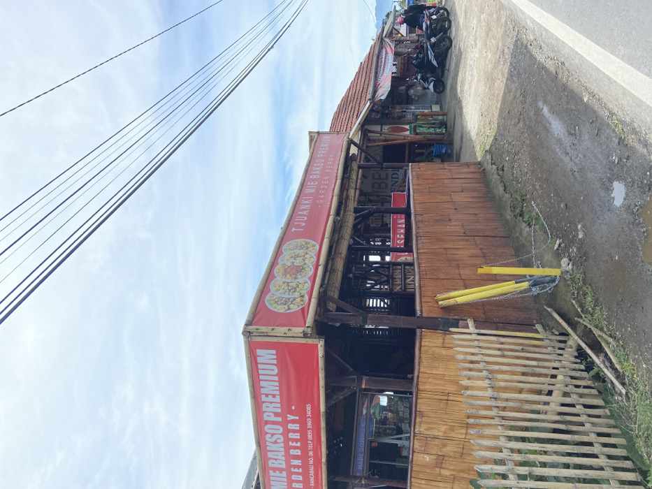 The image shows a roadside restaurant in Rancabali, Indonesia (latitude: -7.122, longitude: 107.428). The restaurant has a red sign that reads "Tjuaniki Mie Bakso Premium - Garden Berry". There is a wooden fence, a chain, and a sign on a pole that reads "Made in China." There is a building to the right of the restaurant. Several motorcycles are parked in front of the building. The sky is clear and the weather is sunny. There are multiple power lines running across the sky. A wooden structure supports the sign. The restaurant appears to be open for business. The road in front of the restaurant is paved with gravel.