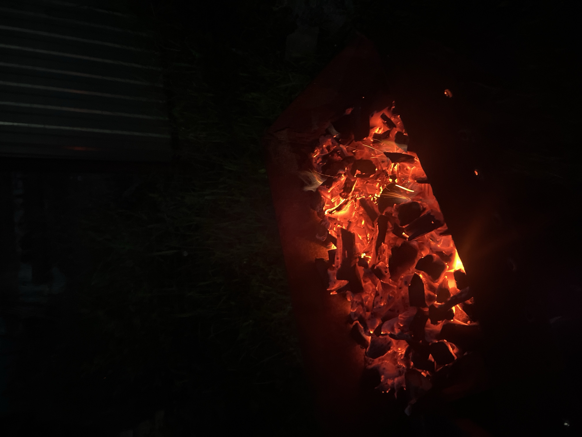 The image displays a nighttime outdoor scene. A rectangular, dark-colored container, likely metallic, occupies the lower central portion of the frame, filled with intensely glowing red and orange charcoal embers. Small, bright sparks and faint yellow-orange flames are visible emanating from the burning material. The surface of the embers is uneven, indicating individual pieces of charcoal.
Surrounding the container is dark, indistinct ground cover consistent with grass or low foliage. In the upper right background, a vertical corrugated metal fence or wall is partially visible, reflecting some light, though its details are obscured by darkness. The remainder of the background is largely black, with no discernible objects or features. There are no individuals present in the scene, and no specific actions or interactions are depicted beyond the burning charcoal. The image itself does not contain specific visual cues to identify its location as Chaikovsky, Russia.