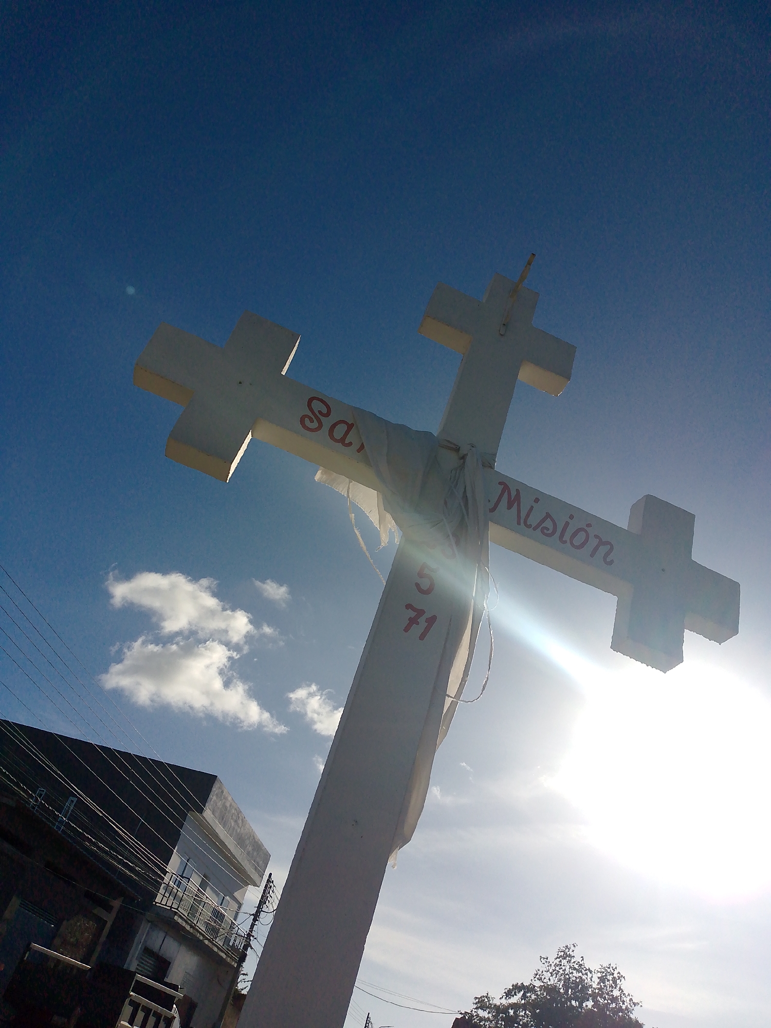 An outdoor scene in Juan Griego, Venezuela, depicts a large white cross viewed from a low angle against a bright blue sky with scattered white cumulus clouds. The substantial cross, likely constructed from concrete or stucco, features red painted lettering: "San" on the left horizontal arm, "Misión" on the right horizontal arm, and "5 1971" vertically below the cross-section. White fabric strips are draped and tied around the intersection of the cross's arms. The sun is positioned in the lower right quadrant, producing a strong lens flare that brightens the lower right portion of the sky and illuminates part of the cross. In the lower-left background, a multi-story building with dark upper sections and balconies is partially visible. Multiple utility wires stretch horizontally across the frame in front of this building. A leafy green tree canopy is visible at the bottom center edge of the image. No individuals or specific actions are discernible.