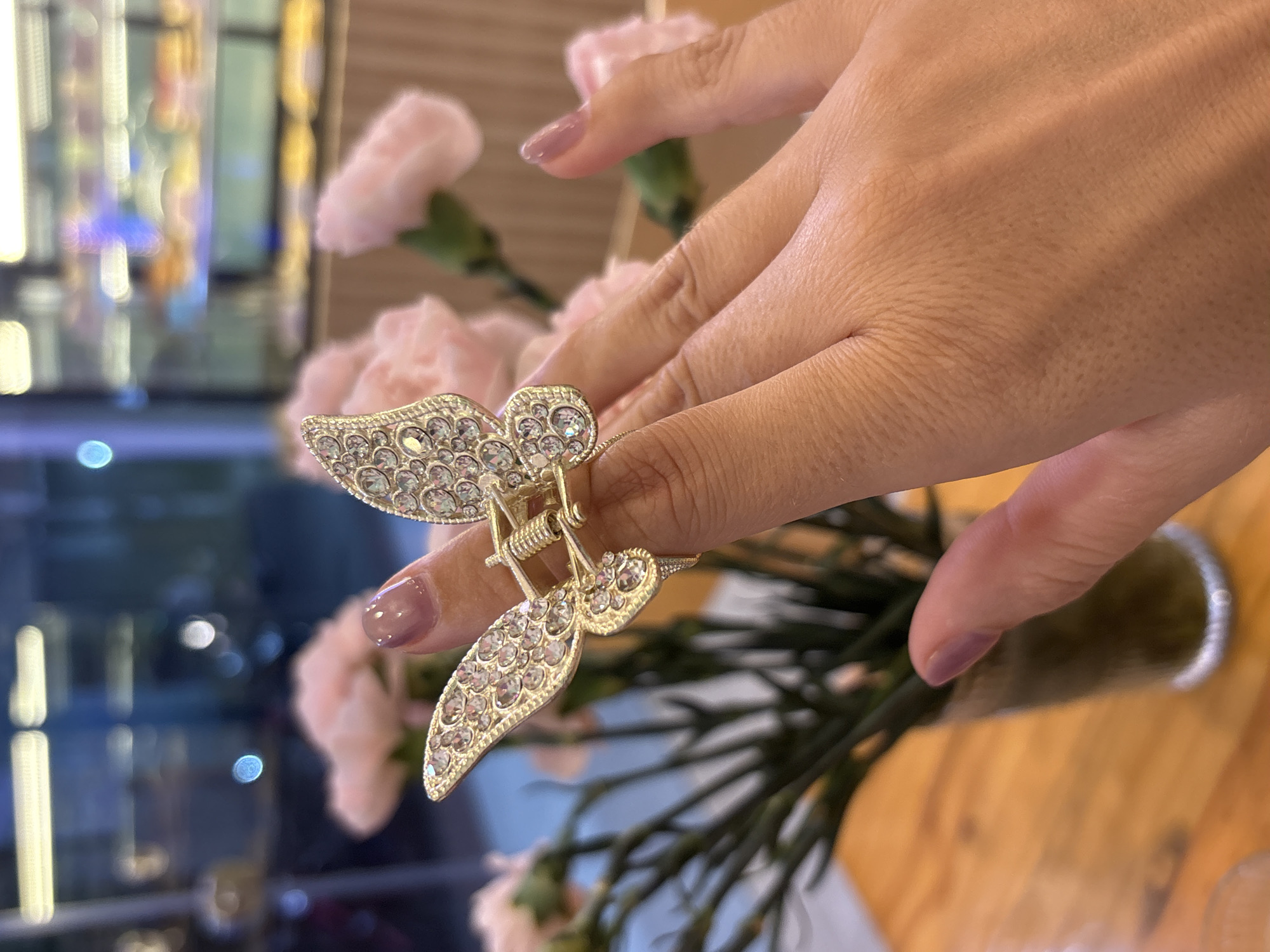 A close-up shows a woman's hand adorned with a gold-toned hair clip shaped like a butterfly, embellished with numerous small, clear stones. The clip is clasped around the ring finger of the hand. The woman has painted her nails a light purple color. The background features a bouquet of light pink carnations in a vase, set against a blurred backdrop of what appears to be an interior space with lighting fixtures. The scene is likely in a city, and the location might be Bac Nha Trang Ward, Vietnam. The table appears to be wooden.