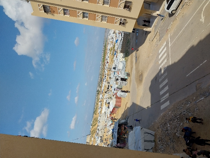 An elevated view captures a street scene in Khan Yunis, Palestinian Territory, flanked by permanent buildings and transitioning into a large temporary settlement. The foreground features a paved asphalt road with a visible white pedestrian crossing. To the left, a dirt embankment hosts a white tent structure and a group of approximately 5-7 individuals, including children, sitting or standing. A child in a blue shirt is running across the dirt area further left. To the right, a multi-story, light-brown building with decorative window grates and a shaded ground-level entrance stands. A white sedan is parked on the street adjacent to this building, with a lone individual in a blue shirt standing nearby. Two additional individuals in dark clothing are partially visible under the building's shaded entrance.
Beyond the road, the mid-ground and background reveal a sprawling camp composed of numerous closely packed temporary shelters constructed from white, grey, and brown tarpaulins and other makeshift materials, extending across a dusty, uneven landscape towards the horizon. Some small commercial stalls or shelters are discernible within the camp area. The sky above is clear blue with scattered white cumulus clouds, casting long shadows across the road and dirt areas, indicating a sunny day. A person in a red shirt is visible deeper within the camp area.