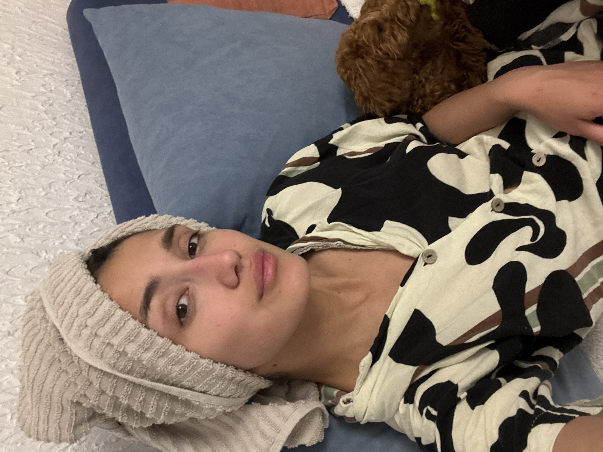 The media file captures a young woman lying relaxed on a bed, looking directly at the camera with a neutral expression. A light beige, ribbed towel is wrapped around her head.
She is wearing a short-sleeved, button-up shirt with a distinctive abstract pattern of black, white, and brown shapes, and light-colored buttons are visible. Her right arm is bent and rests on the bed.
The bed is covered with a white, textured bedspread or sheet. She is resting against a light blue pillow, with a darker blue pillow visible behind it. Partially visible near her head, on the bed and next to the blue pillow, is a brown, fluffy object, which appears to be a small dog.
The setting is an indoor space, likely a bedroom, characterized by soft lighting. The activity depicts the woman relaxing, possibly after a shower. There is no visible text in the image. The location context for this scene is Sutomore, Montenegro.