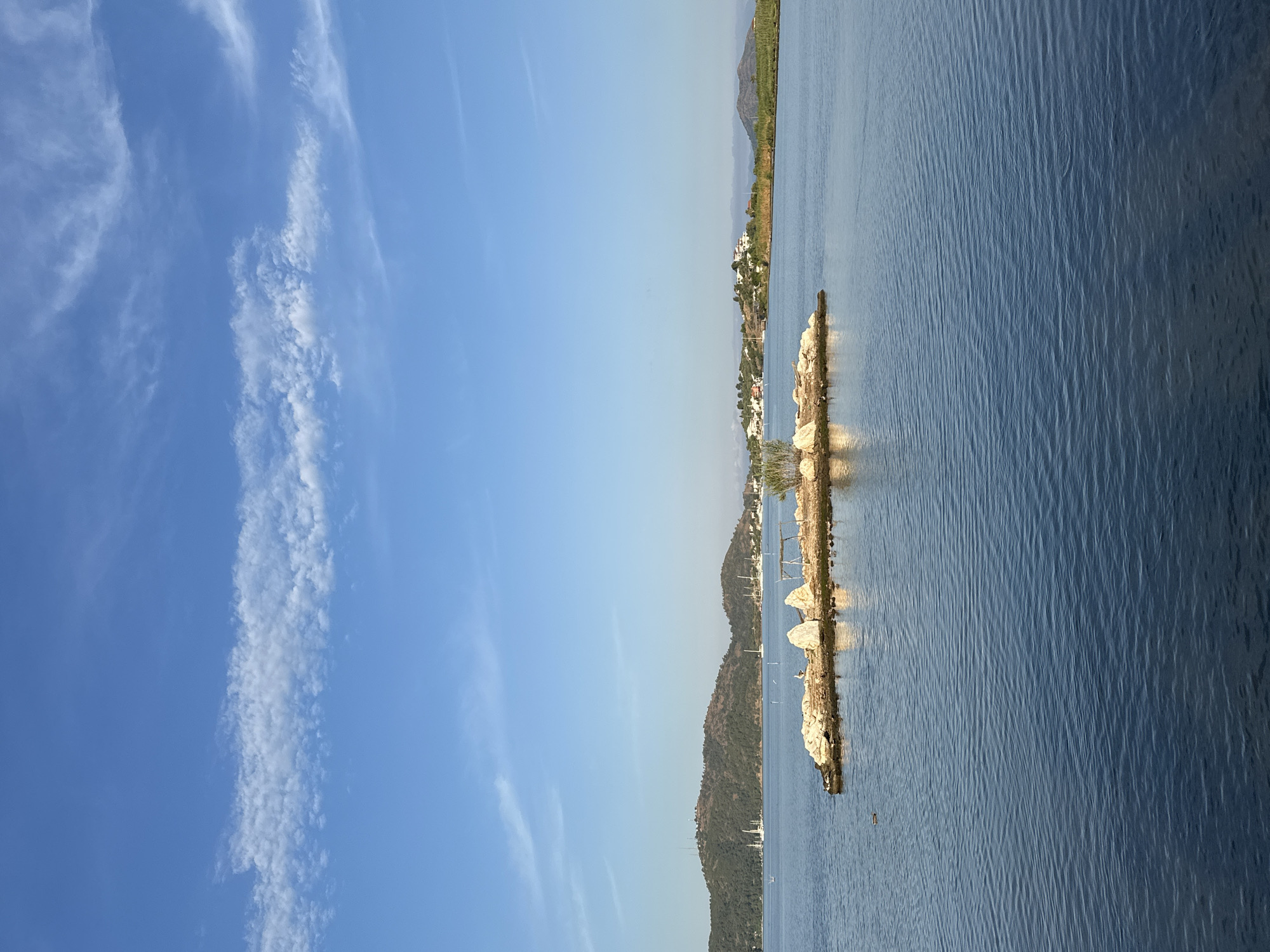 The image captures a serene coastal landscape under a bright, clear blue sky. When correctly oriented, the horizon line runs horizontally, separating the sky on the upper left from the water and distant land on the lower right. The sky is largely clear, with several long, wispy white clouds stretching across it.
The main body of water, likely a sea or a large bay, is calm with visible ripples on its surface, reflecting the blue of the sky. From the right side of the frame, a natural or artificial rocky outcrop extends into the water, appearing as a line of light-colored stones.
In the mid-ground and background, verdant hills and mountains rise from the water's edge. Scattered along the hillsides are clusters of white buildings, indicative of a settlement or coastal town. A few small boats or yachts are visible in the water near the distant shores.
The scene depicts a sunny daytime, likely late morning or early afternoon, characterized by bright light and excellent visibility. The overall atmosphere is peaceful and tranquil, consistent with a picturesque coastal location such as Fethiye, Turkey. There are no people visible, nor is any text present in the image.