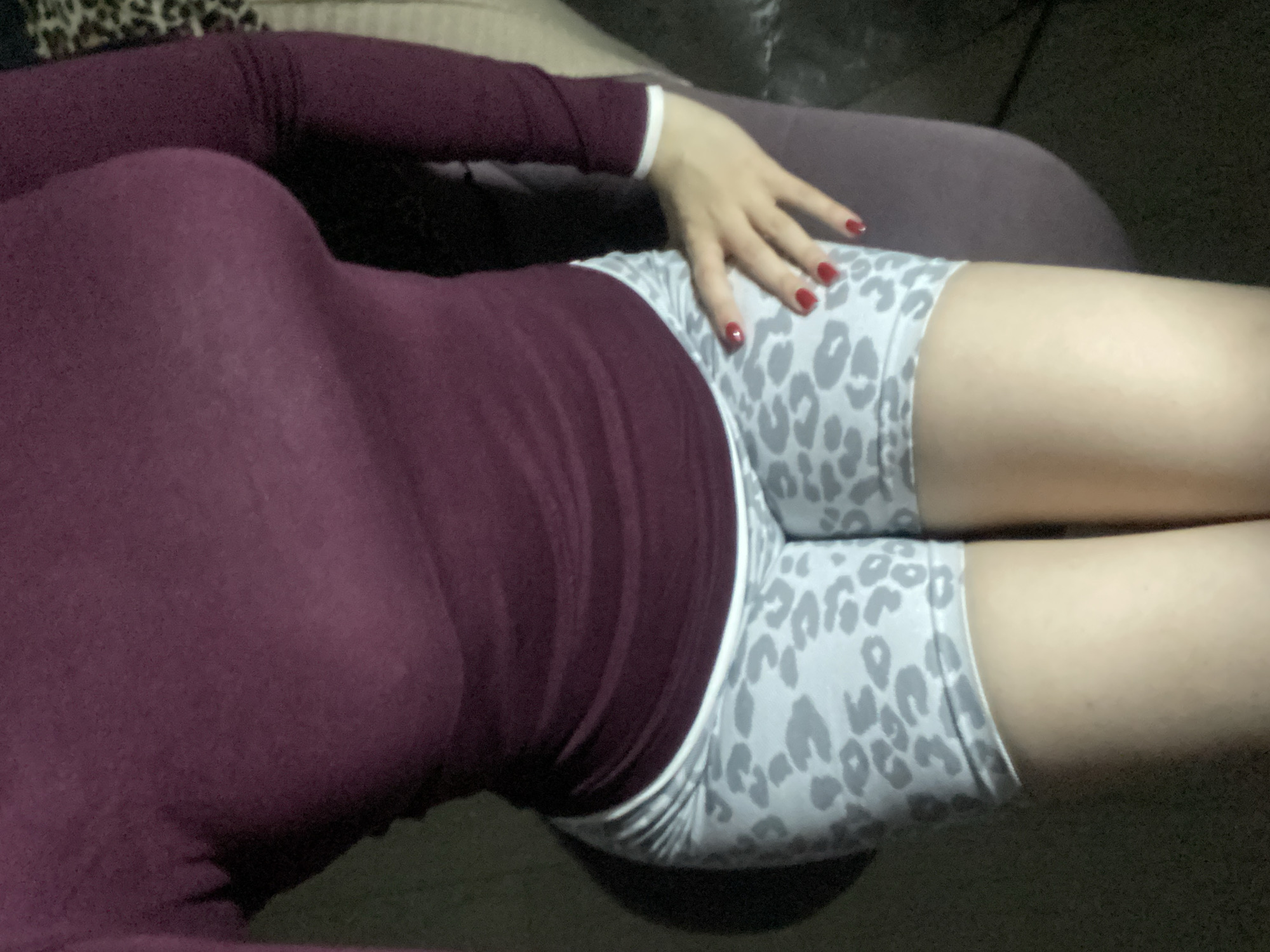 A medium-angle, eye-level shot shows a person reclining indoors in low light. The image is oriented with the person's body spanning diagonally from the lower left to the upper right. Only the torso, upper thighs, and part of one arm are visible, suggesting a close-up, cropped view.
The individual is wearing a form-fitting, long-sleeved top or dress in a dark plum or maroon color. The fabric appears soft and slightly sheer, revealing the general contours of the upper body. Below the waist, they wear light-colored shorts with a grey leopard print pattern, possibly made of a stretchy material like biker shorts.
One hand, with neatly painted red nail polish, rests gently on the person's upper thigh, overlapping the top of the leopard print shorts. The skin tone is light. The person's legs extend out of the frame towards the right.
The background is indistinct and dark, suggesting an indoor setting, possibly a room with muted lighting or taken during the evening. A soft, dark fabric surface, likely a sofa or bed, is visible behind and beneath the person. The lighting is dim, creating some soft shadows and highlighting certain features like the hand and parts of the legs. No specific activity is clearly depicted other than posing for the photograph in a relaxed posture. There is no visible text in the image.
