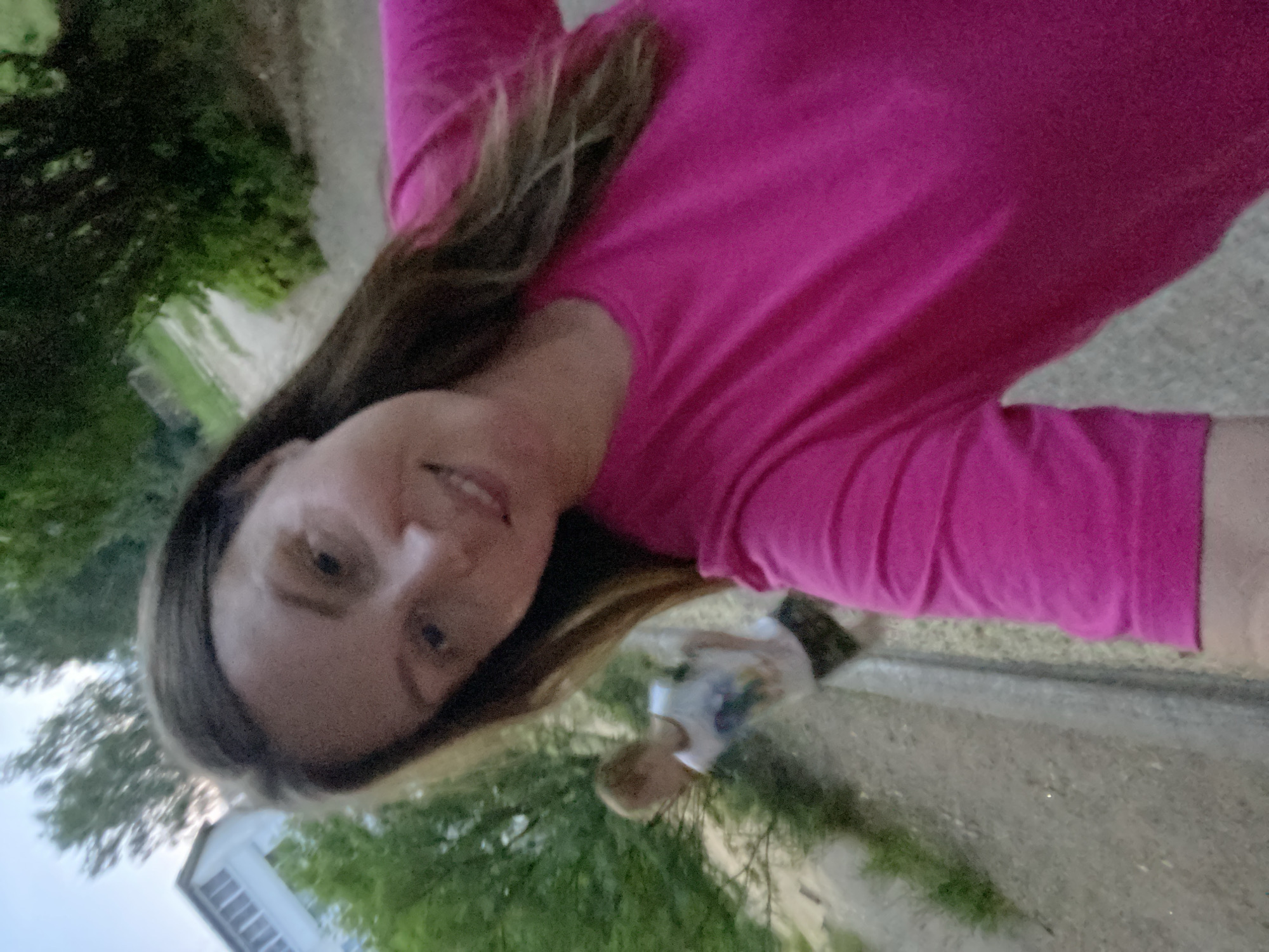 This is a selfie taken outdoors by a woman wearing a bright pink long-sleeved shirt. Her face is close to the camera, and she is smiling. Her hair is long and brown. In the background, there is greenery, suggesting a natural setting. A small child is visible in the background, facing away from the camera. Further in the background, a white building with a pitched roof is partially visible. The image appears to be taken during the daytime, possibly in the late afternoon or early evening, given the lighting conditions. The overall mood of the image is casual and joyful.