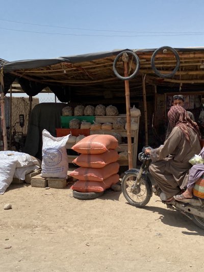A daytime shot depicts a roadside vendor in Sard-E 'olya, Afghanistan. The vendor's stall is constructed with a makeshift canopy overhead, supported by wooden posts, with two tires mounted as decoration. Rows of goods are visible on shelves behind a stack of four reddish-orange sacks on a tire. A white sack with blue writing sits on concrete blocks next to additional white bags. A man in a brown robe and headscarf sits on a motorcycle, appearing to be engaging with another man partially visible behind the vendor's display. A second person is sitting behind the man on the motorcycle. The scene is situated on a dirt surface.