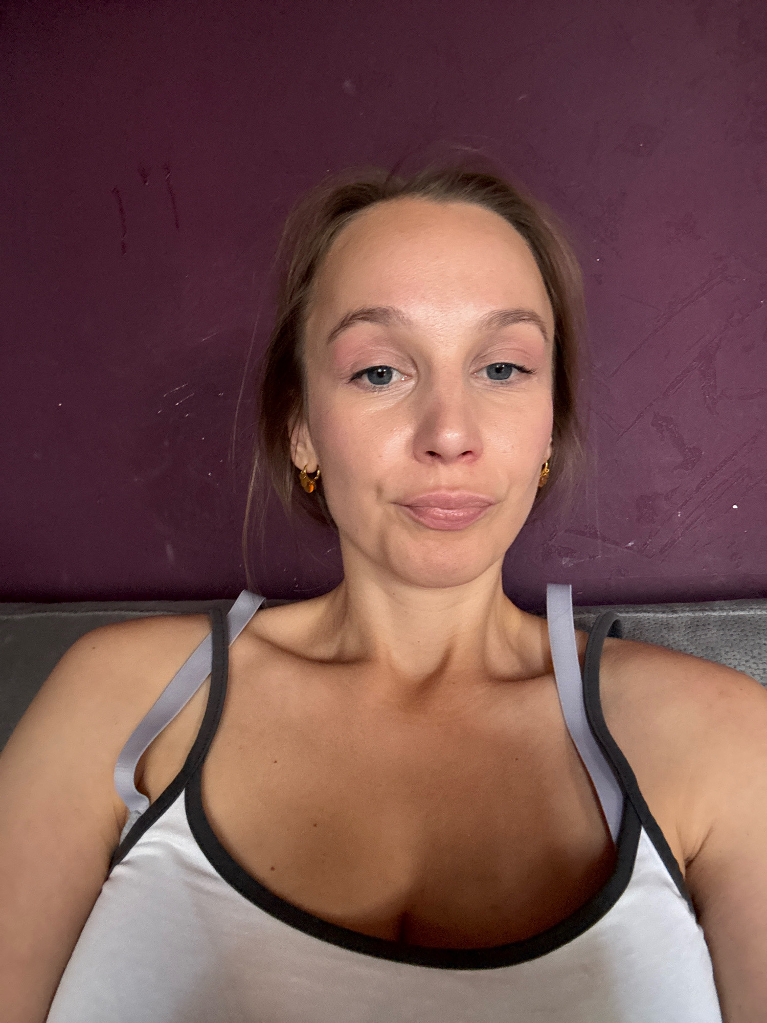 The image shows a selfie of a woman taken indoors, with a plain purple wall in the background. The woman is looking directly at the camera with a slight smile. She is wearing a white tank top with black and gray trim. She has blonde hair and blue eyes. There are some faint marks on the wall that resemble scratches or smudges. The lighting appears to be natural, coming from the front and slightly from above. The overall impression is of a casual, unposed photograph. There is no information available to suggest the location is in Überherrn, Germany.