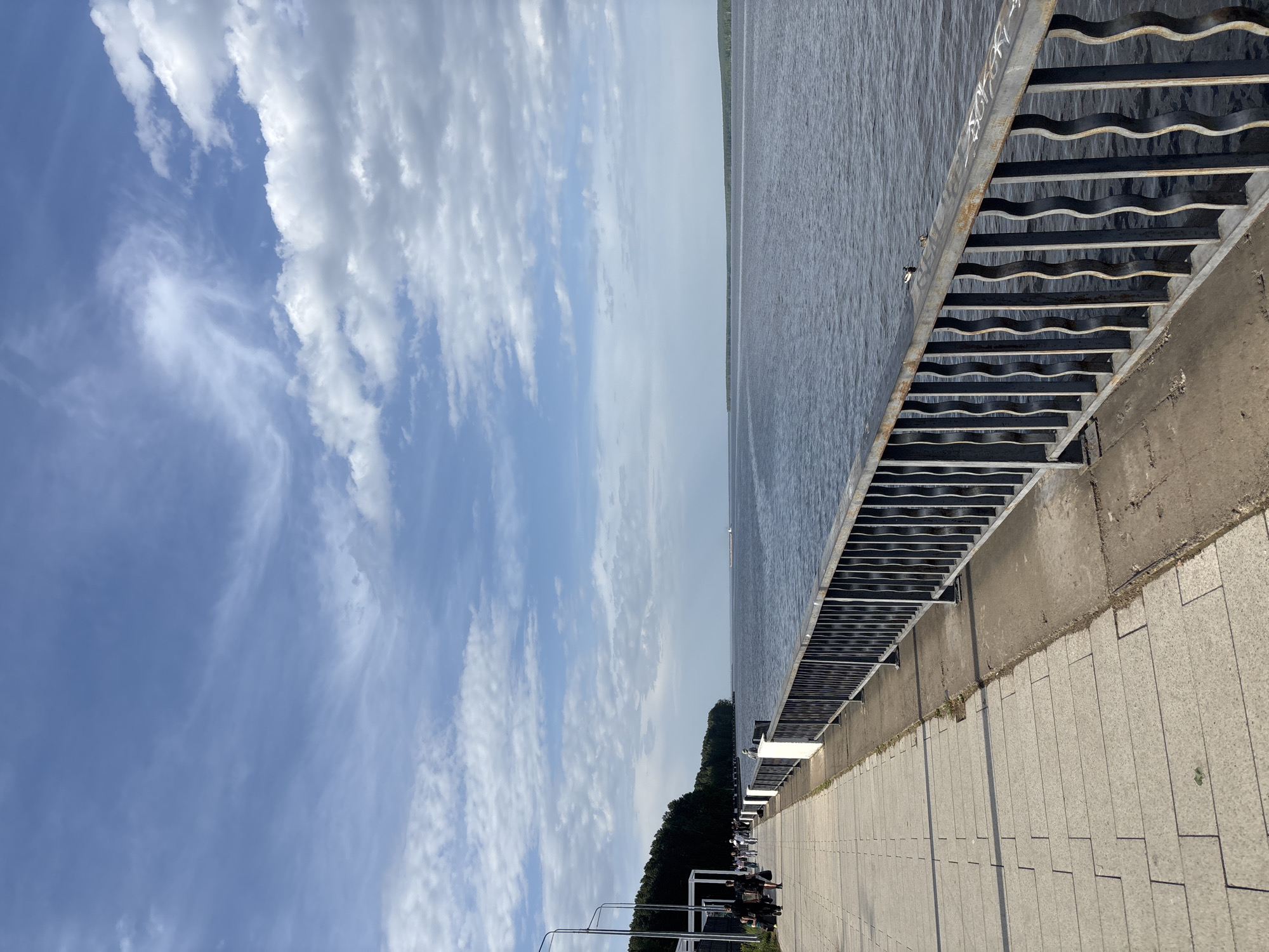The image depicts a waterfront scene. On the right side, there is a raised concrete walkway or promenade paved with rectangular stones. A metal railing runs along the water's edge. The water itself is dark and appears calm. Across the water, a strip of land is visible. The left side of the image shows a blue sky with scattered white clouds. Some trees and objects are on the bottom-right of the image.