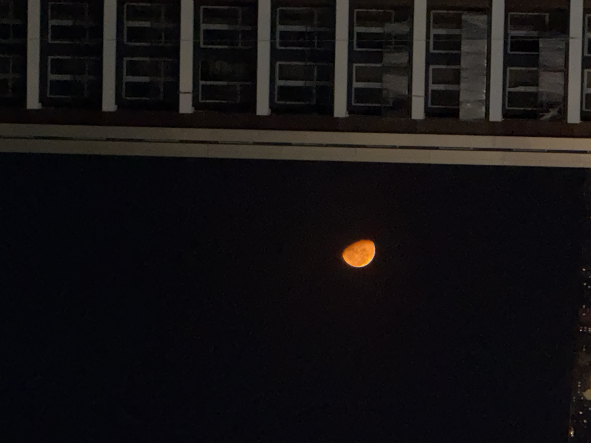 An orange crescent moon hangs in the dark night sky over Tbilisi, Georgia, partially visible to the left of a tall, multi-story building. The building, with its dark facade and rows of windows and balconies, occupies the right side of the frame. The moon is positioned in the upper left quadrant, with the building extending from the upper right down to the bottom edge. A faint line of city lights is visible at the very bottom of the image.