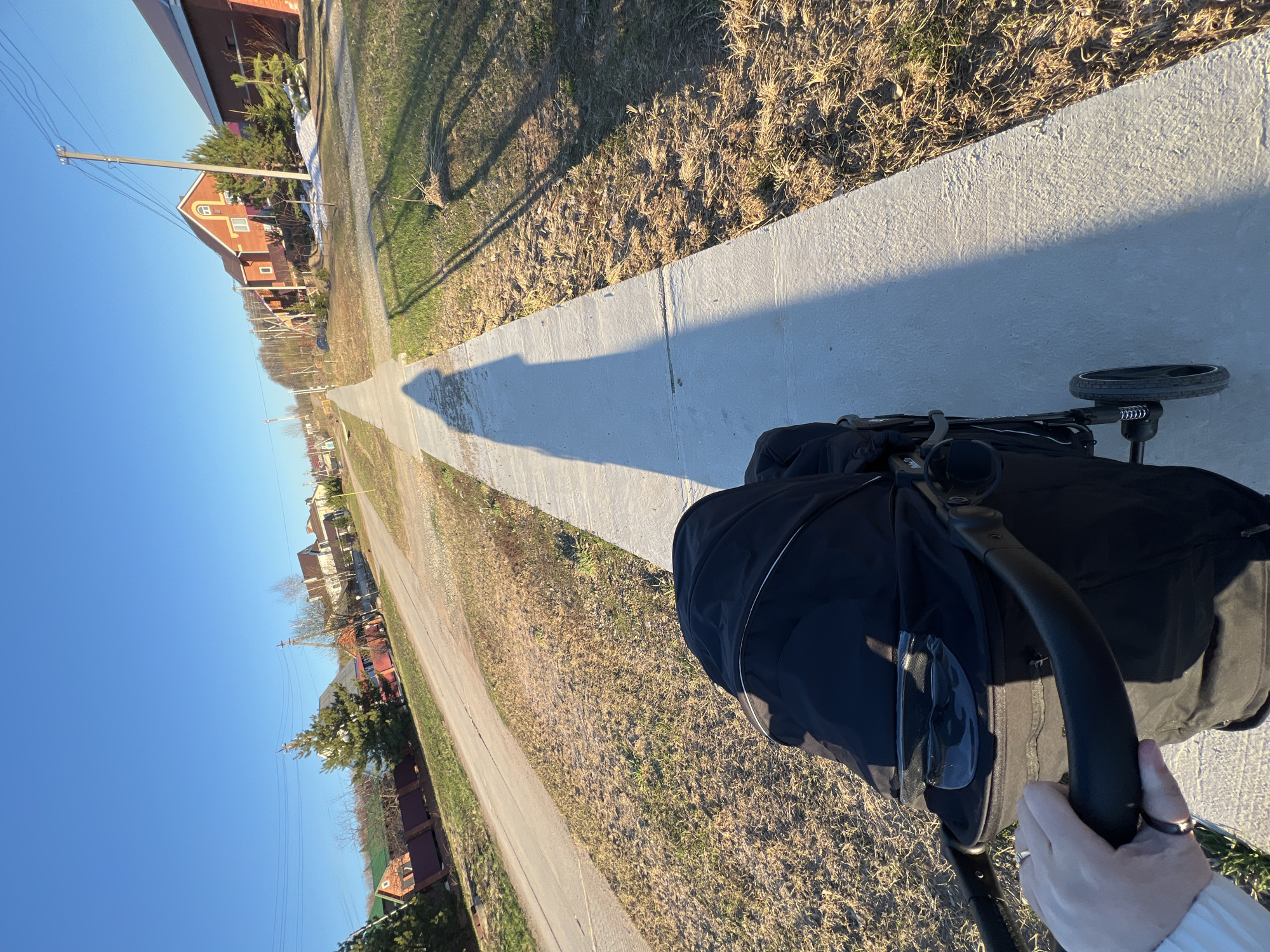 A view of Noviy, Russia, is captured from a first-person perspective behind a black stroller on a sunny day. The stroller is being pushed along a concrete sidewalk. A line of houses borders the sidewalk, stretching into the distance with dry grass and budding vegetation in between. Overhead power lines are visible against the clear blue sky. The shadows of trees or poles are cast onto the sidewalk. The hand of the person pushing the stroller is visible holding the handle.