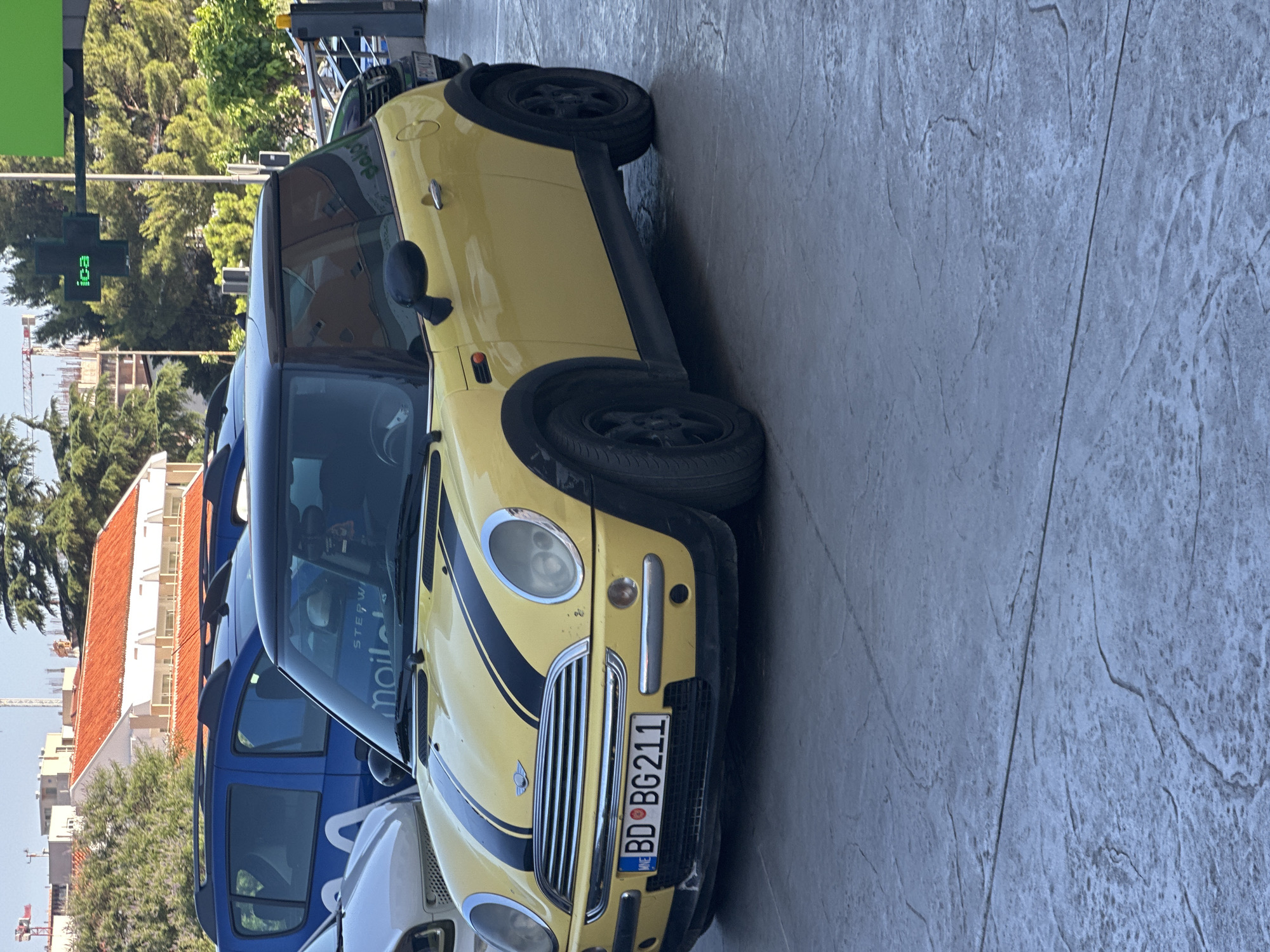A yellow Mini Cooper with black racing stripes on the hood is parked on a stone-patterned sidewalk. The car has a black roof and black wheel arches. The license plate reads "BD BG211". In the background, a blue car is parked behind the Mini Cooper, and a building with an orange tiled roof and construction cranes are visible. A green sign with the word "ica" in white letters is mounted on a pole to the right of the Mini Cooper. The scene appears to be an urban environment, likely in Budva, Montenegro, given the license plate format. The lighting suggests it is daytime, with sunlight casting shadows.