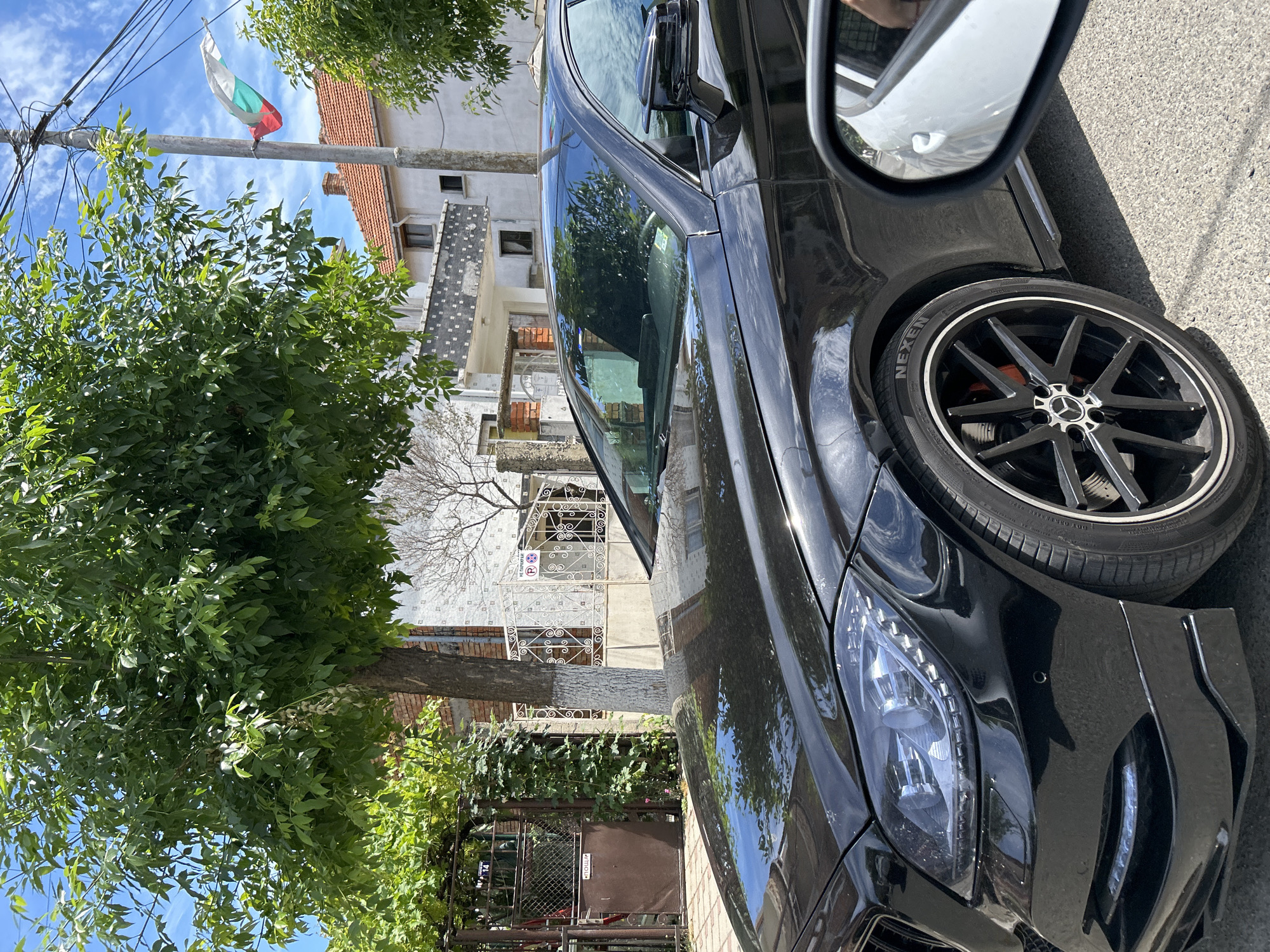 A black Mercedes-Benz sedan is parked on a street in Aheloy, Bulgaria. The car is facing the right of the frame, with its front passenger side and wheel prominently displayed. The vehicle has a sleek, modern design, with LED headlights and a sporty black rim. The tire has "NEXEN" written on it. A side mirror reflects a bit of the surroundings, including some greenery.
Behind the car, a residential building with a red-tiled roof and light-colored walls is visible. The architecture suggests a European setting. A Bulgarian flag, with its white, green, and red stripes, is flying from a utility pole in the background, adding a clear location cue. Lush green trees with dense foliage flank the street, providing shade and a natural element to the scene. The sky is bright blue with some scattered clouds, indicating a clear and sunny day. The time appears to be daytime. No people are visible in the image. The overall scene is calm and typical of a residential street in a town.