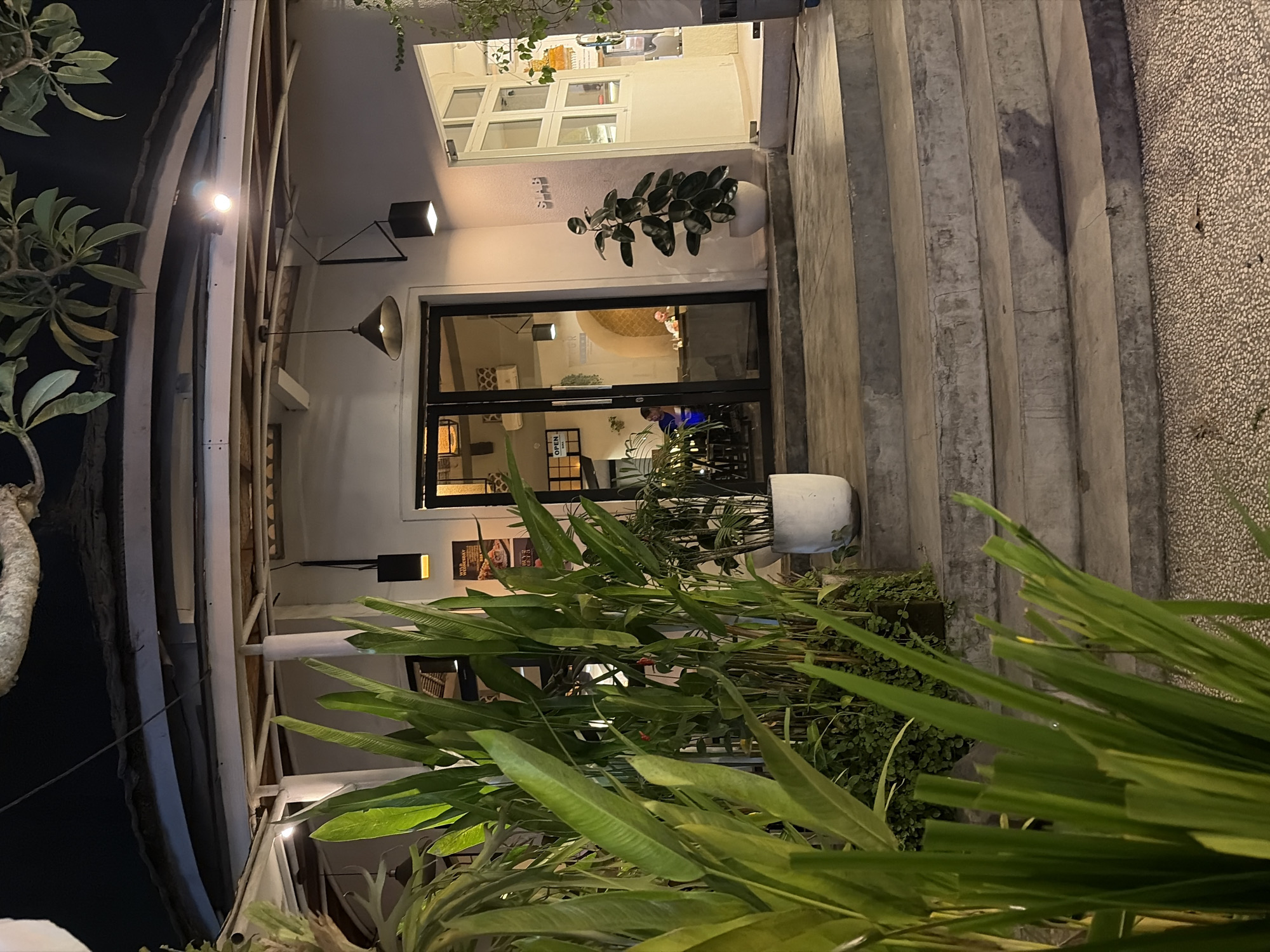 The image depicts the entrance of a restaurant or cafe at night in the Ubud District, Indonesia. The building has a modern architectural style with white walls and a sloping roof covered in thatch or a similar material. Large glass doors lead into the establishment, revealing people seated at tables inside. Several potted plants are strategically placed around the entrance, including tall, leafy plants in the foreground and a rubber plant in a white pot to the right. A set of concrete stairs leads up to the entrance, with a textured pebble-like material forming the ground surface. Lighting fixtures, including pendant lamps and wall-mounted lights, illuminate the exterior. The overall atmosphere suggests a contemporary dining establishment integrated with natural elements.