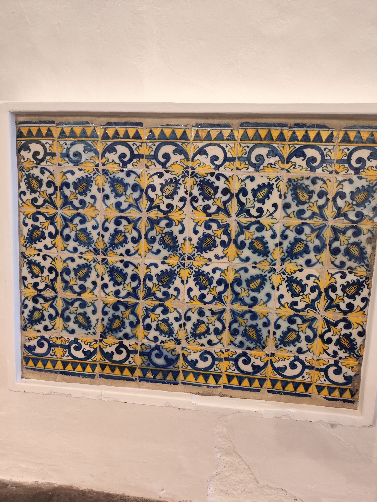 The image presents a rectangular panel of traditional Portuguese azulejo tiles embedded within a textured white wall. The dominant colors of the tiles are a vibrant dark blue and bright yellow against a white or off-white background.
The main body of the tile panel features a repeating, interlocking floral or botanical pattern. Each individual square tile typically contains a quarter of a larger design, where stylized dark blue elements, possibly representing leaves or petals, are intricately detailed with yellow accents and radiating tendrils. When four tiles meet, they complete a symmetrical, star-like floral motif.
Along the upper and lower edges of the central tile pattern, there are distinct border tiles. These border sections display a pattern of blue scrollwork, topped by a continuous band of small yellow triangles, creating a saw-tooth or zigzag effect.
The entire azulejo panel is encased by a clean, smooth white frame, which contrasts with the slightly rougher, stucco-like texture of the surrounding white wall. Some tiles show minor signs of wear or slight imperfections, suggesting their age and authenticity. The lighting is even, highlighting the intricate details and colors without harsh shadows. Given the provided context, this decorative tile panel is located in Ponta Delgada, Portugal, characteristic of the region's architectural embellishments. There is no visible text in the image.