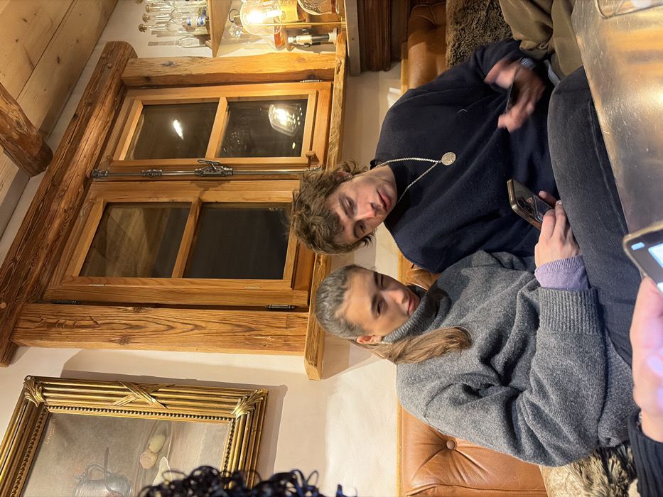 An indoor scene depicts two young adults seated on a brown tufted leather couch, engaged with smartphones, within a rustic establishment consistent with an alpine lodge or restaurant in La Clusaz, France.
The individual on the left is a female with long brown hair tied in a ponytail, wearing a grey turtleneck sweater over a light purple long-sleeved shirt, and dark pants. She holds a dark smartphone horizontally with both hands, looking towards the camera with her left eye partially closed.
The individual on the right is a male with messy light brown hair, wearing a dark blue crew-neck sweater and dark pants. He has a silver-colored chain necklace with a circular pendant. He holds a dark smartphone in his right hand, looking downwards at the screen.
In the foreground, the hands of a third person are visible, also holding a dark smartphone over a reflective surface.
The background features a prominent wooden-framed window with multiple panes and antique metal latches, flanked by thick wooden beams. To the left of the window, a framed painting in an ornate gold frame depicts a still life. To the right, a wooden shelf unit holds several clear glass bottles, and a decorative lamp with a visible filament bulb provides warm ambient light. The interior exhibits extensive use of natural wood, including a visible ceiling beam, reinforcing a traditional alpine chalet aesthetic.