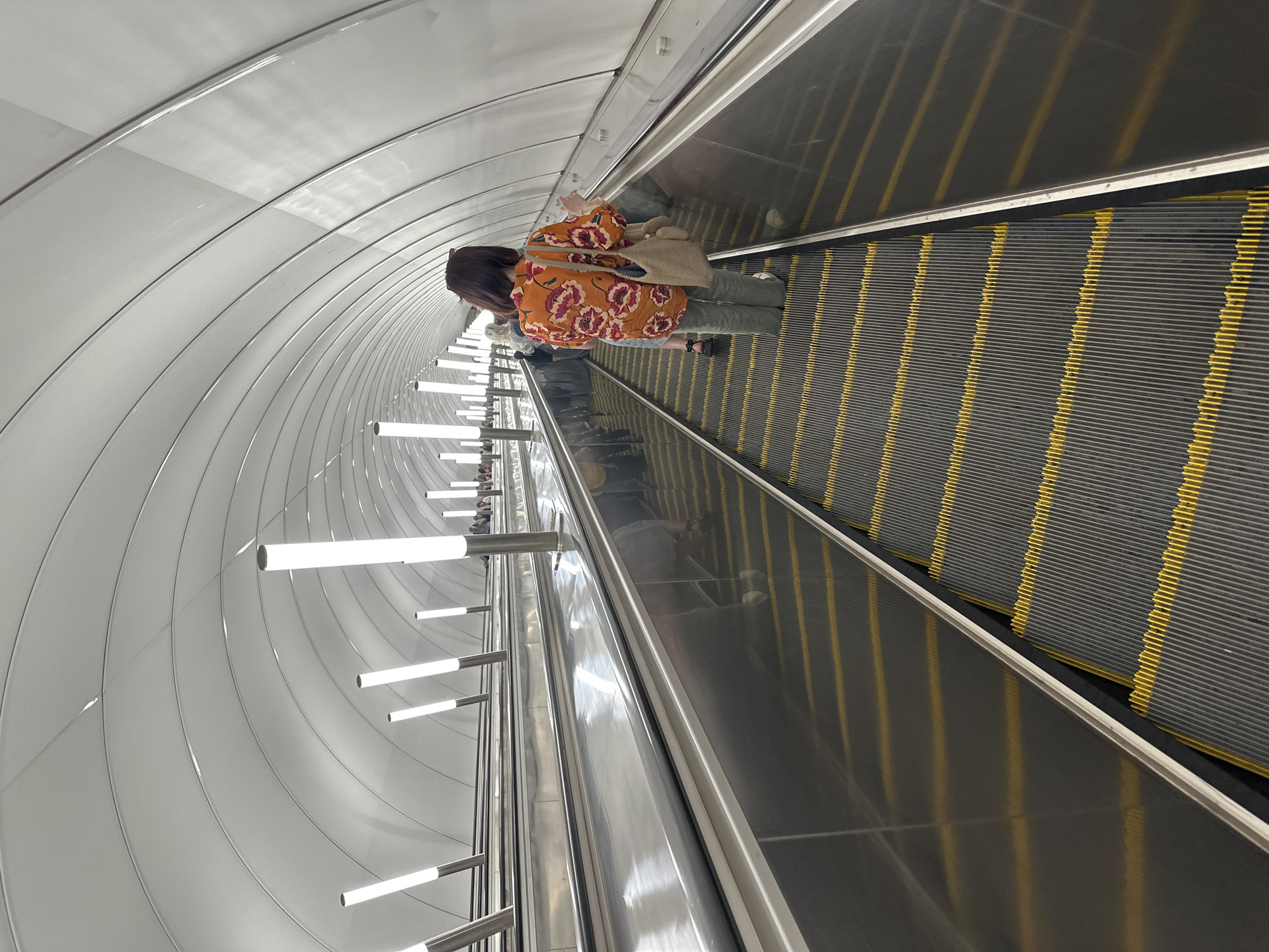 A woman is riding an escalator in a modern, tunnel-like environment. She is seen from behind, facing upwards on the escalator, wearing an orange blouse with a floral pattern and green pants. She carries a light-colored bag slung over her shoulder. The escalator has dark treads with yellow safety lines. The ceiling of the tunnel is a ribbed, white material, curving upwards and forming a wide arch. Linear fluorescent lights are installed along the curve of the ceiling, casting a bright, even illumination. The scene appears to be indoors, likely in a public transportation hub such as a metro station, given the escalator and the architectural style, consistent with some stations in Saint Petersburg, Russia. The lighting suggests it could be daytime or a well-lit underground space. There is no visible text in the image. The overall impression is one of clean, contemporary design.