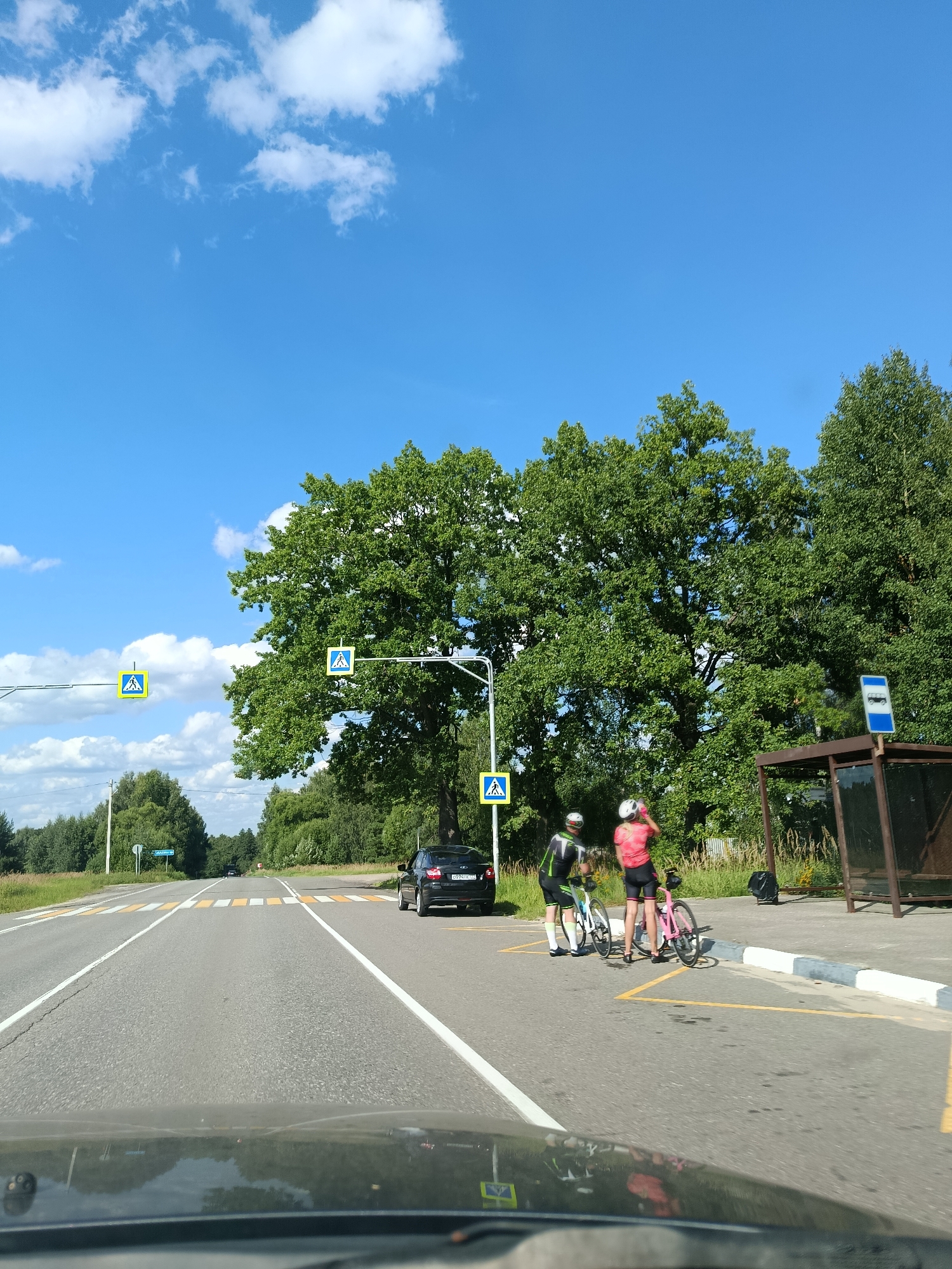 Two individuals, dressed in cycling attire, stand with their bicycles at a pedestrian crossing next to a bus stop. The scene takes place in Novonikolaevka, Russia, on a sunny day with a bright blue sky and scattered white clouds. A paved road with yellow and white pedestrian crossing markings runs through the foreground. A black car is parked to the left of the pedestrian crossing, and a bus stop shelter is visible on the right. Large, green trees line the side of the road, and a forest stretches into the background. The perspective suggests the image was taken from inside a vehicle.